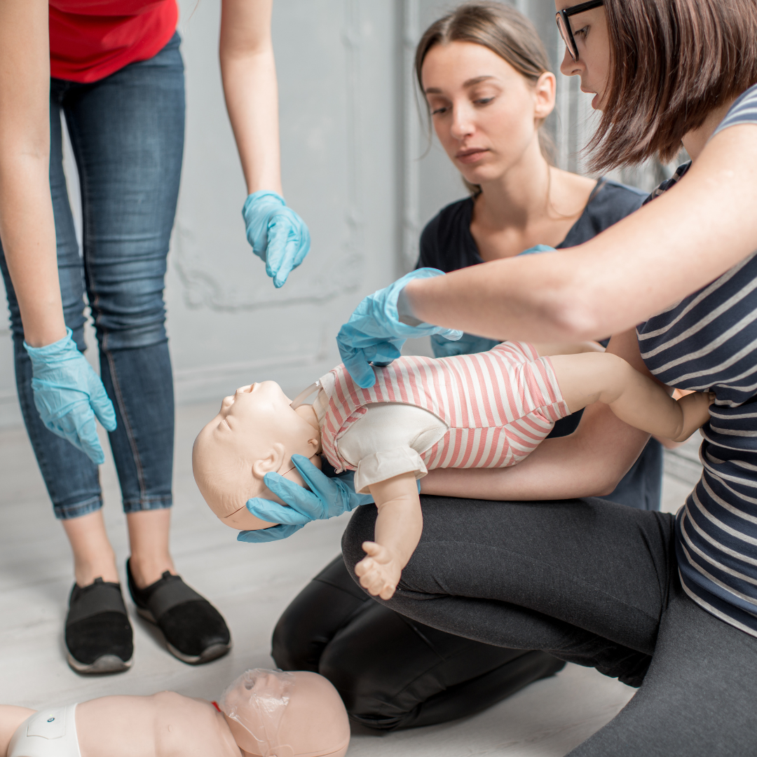 People practicing infant first aid on a baby mannequin, with two women wearing gloves and a third woman supervising.