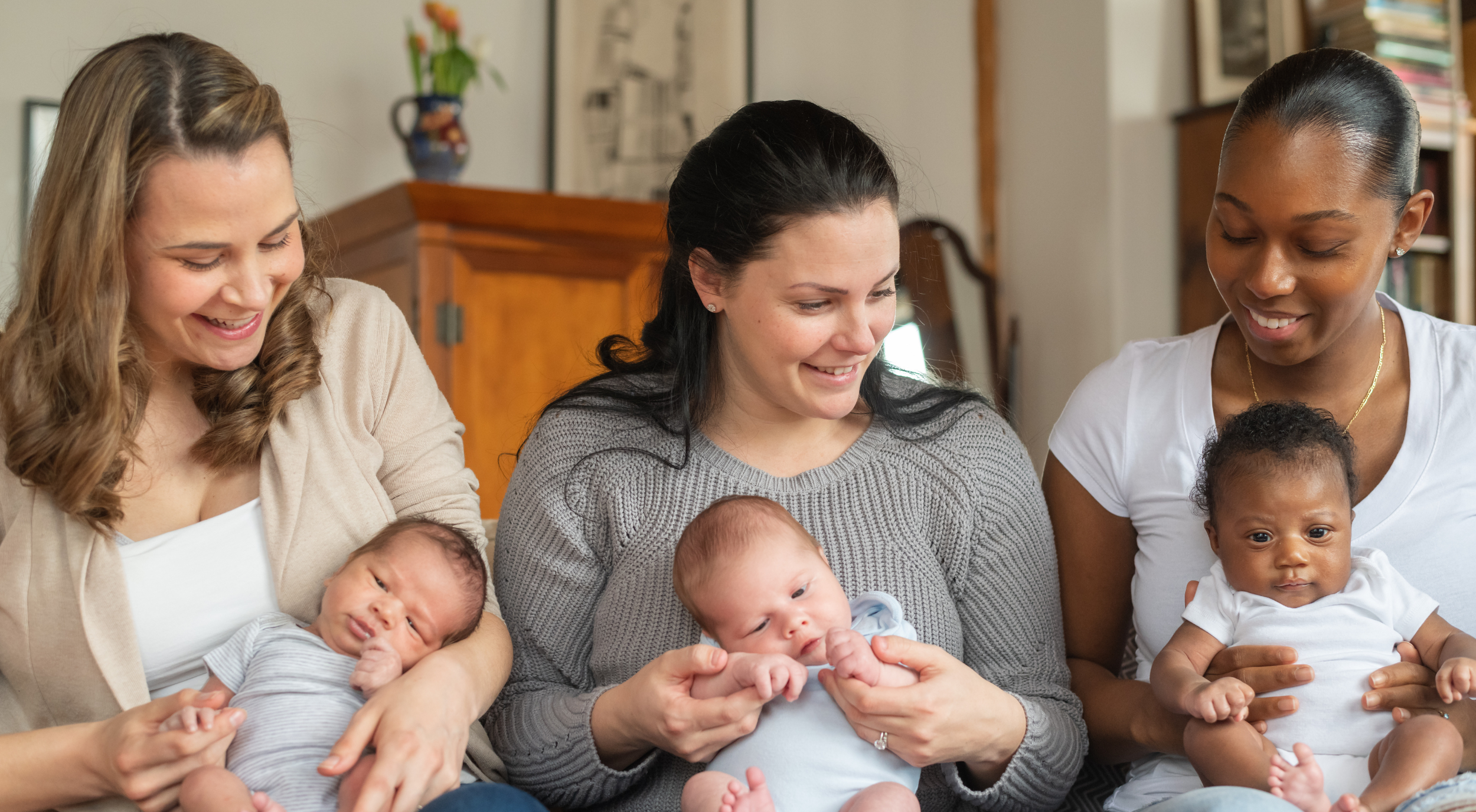 Three women sitting together, each holding a baby, smiling and looking down at their infants in a cozy indoor setting.