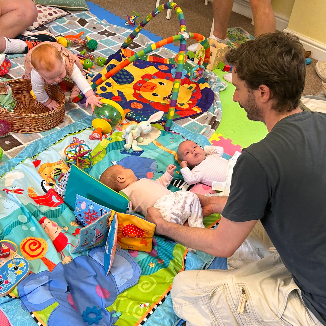 A man is holding two babies lying on colorful play mats with various toys and stuffed animals, while a toddler reaches for toys surrounded by more toys in the background.
