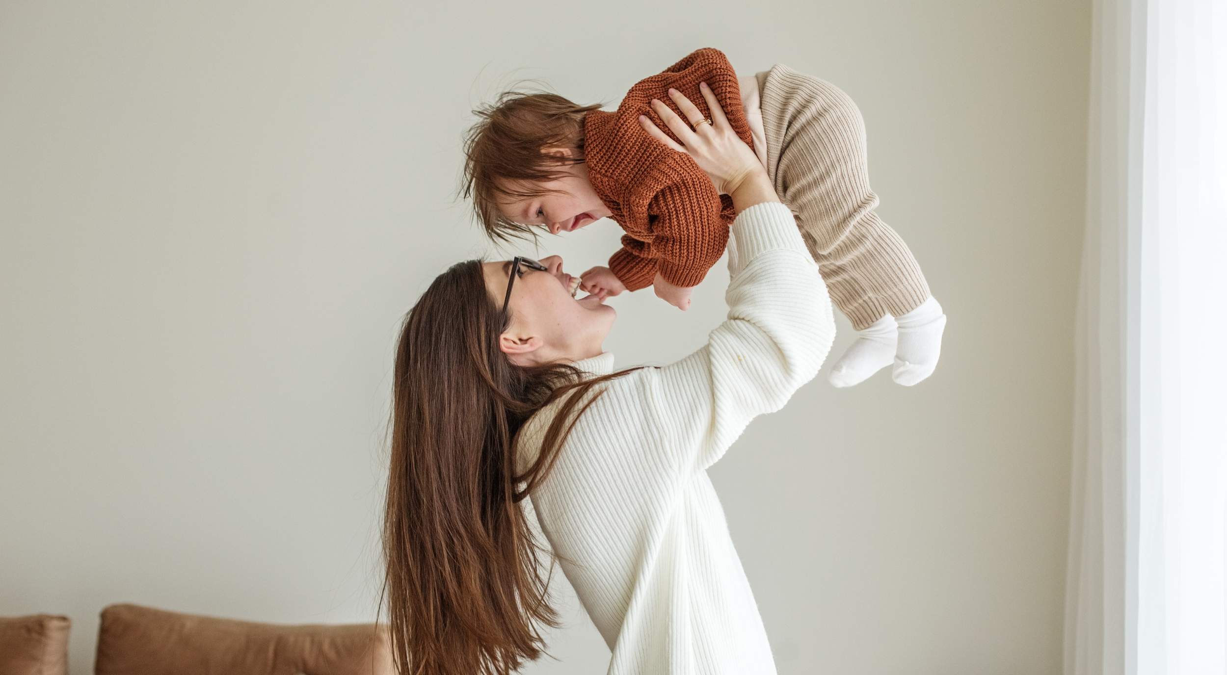 A woman with long brown hair and glasses is lifting a young girl with brown hair and a rust-colored sweater in the air, both smiling and laughing in a room with light-colored walls and curtains.