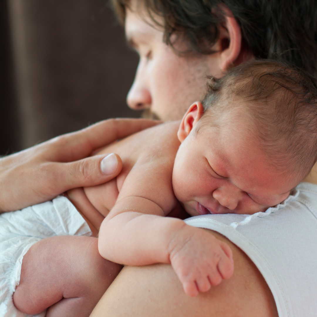 Close-up of a newborn baby sleeping on a man's shoulder, with the man's face gently touching the baby's head.