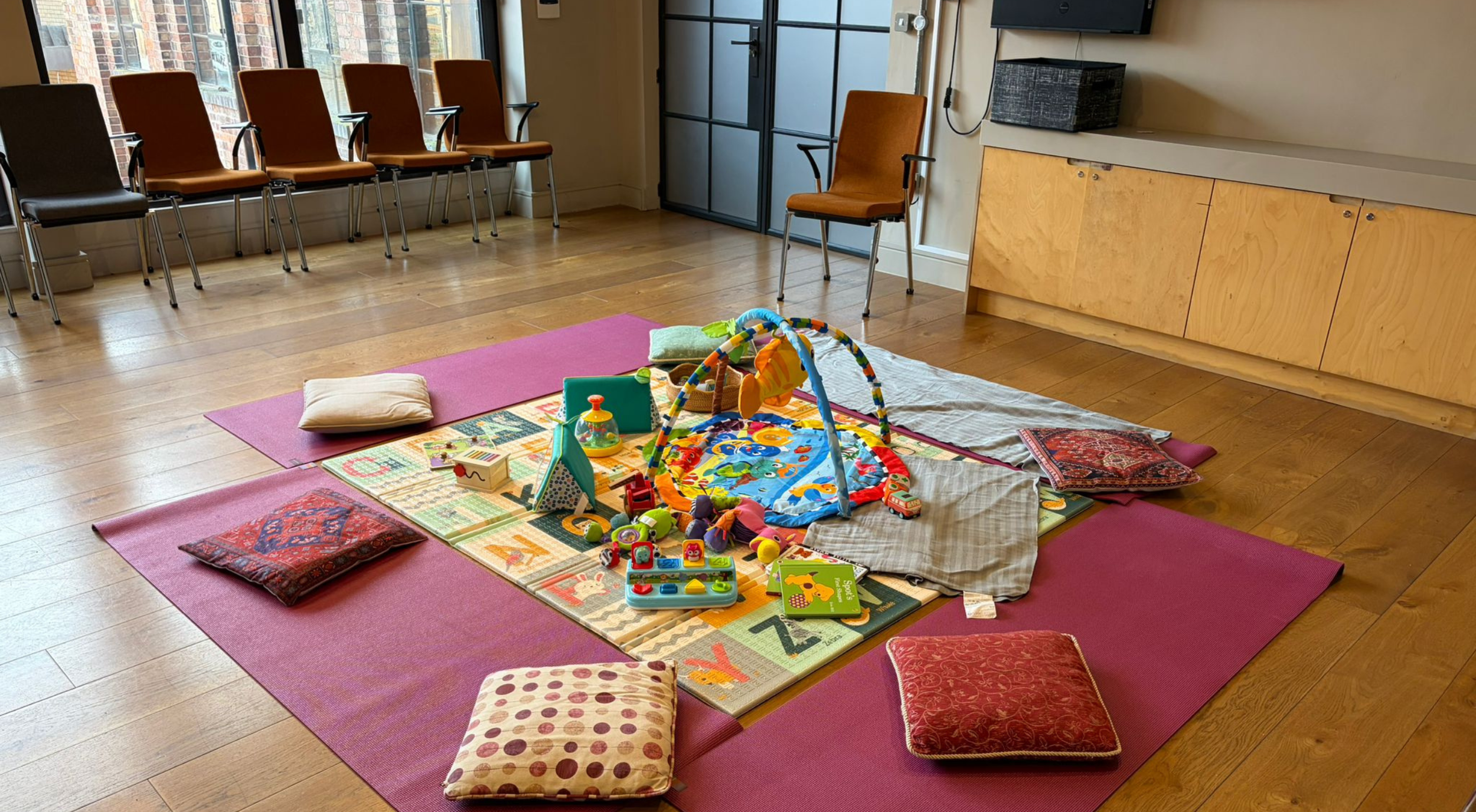 Children's play area with toys and a play mat inside a room with wooden floor, pink mats, and pillows, surrounded by chairs and a cabinet.