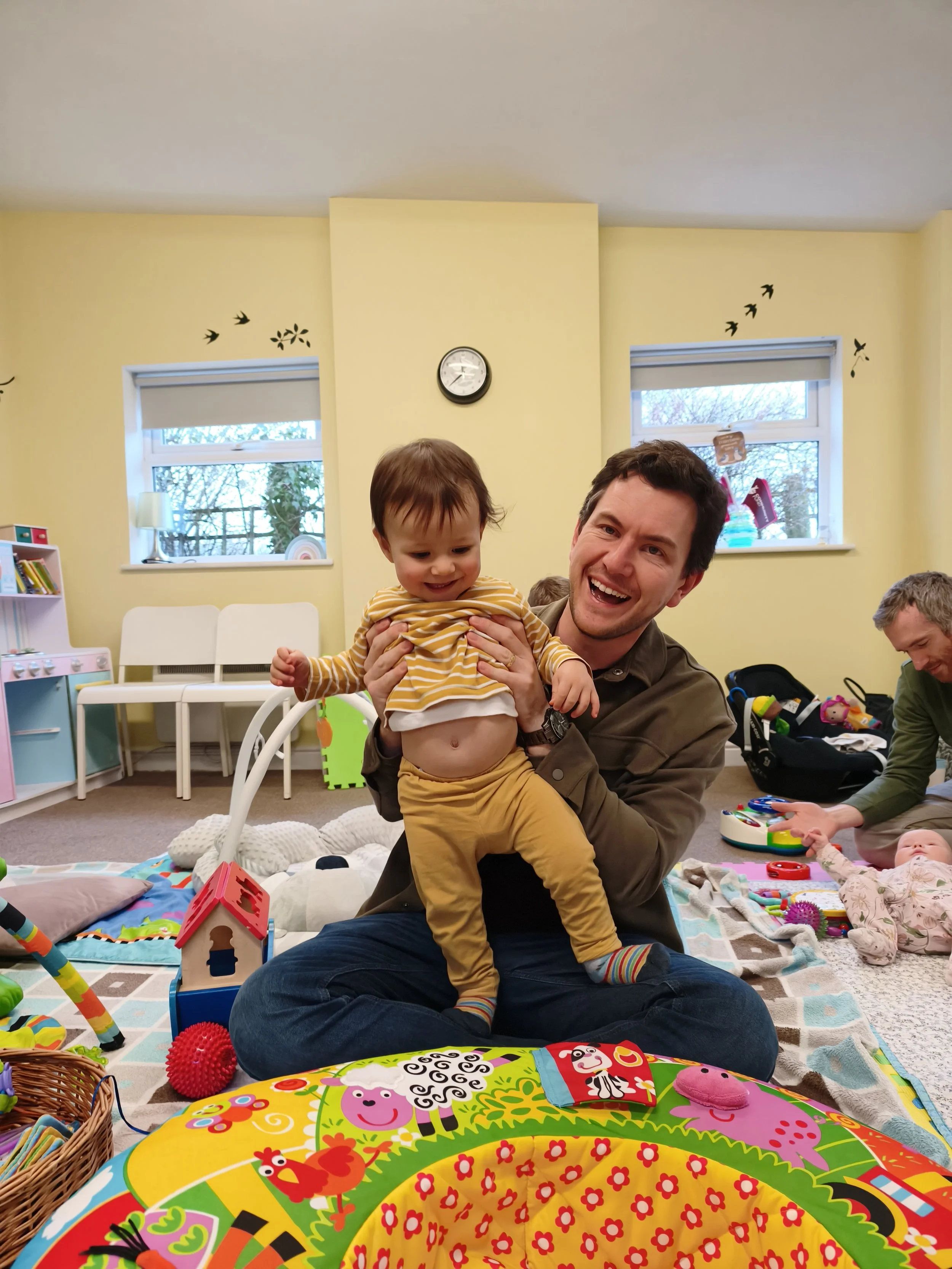 A man is holding a young child with a striped yellow shirt and yellow pants in a room filled with toys, smiling and playing. In the background, another adult is playing with a baby on a blanket. The room has light yellow walls, two windows, and decorative bird decals.