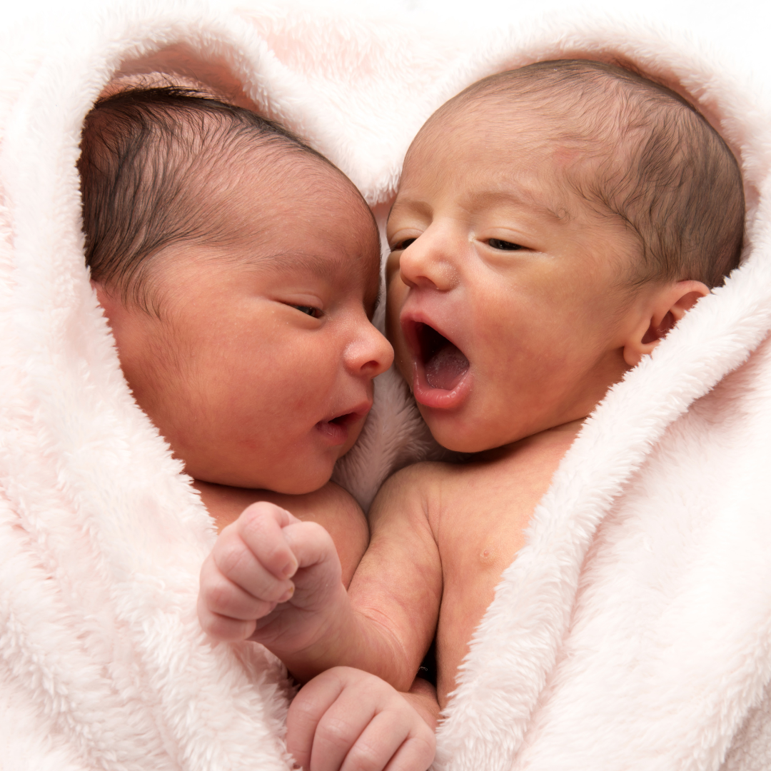 Two newborn babies wrapped in a soft, white blanket, one is yawning and the other appears alert.