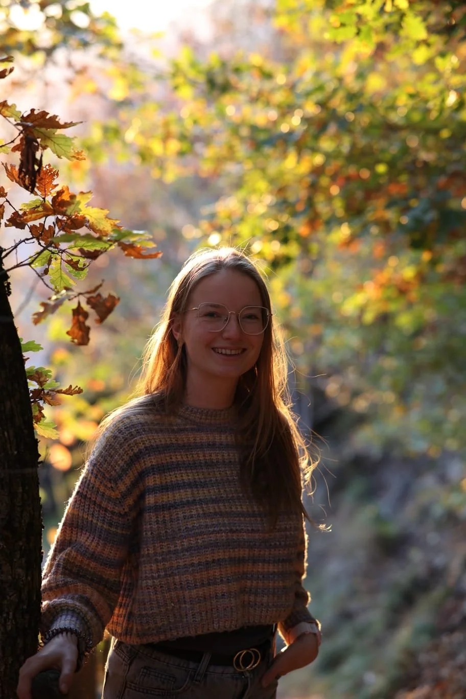 Lächelnde junge Frau mit Brille im Sonnenlicht in einem Baumhain im Herbst, umgeben von bunt gefärbten Blättern.