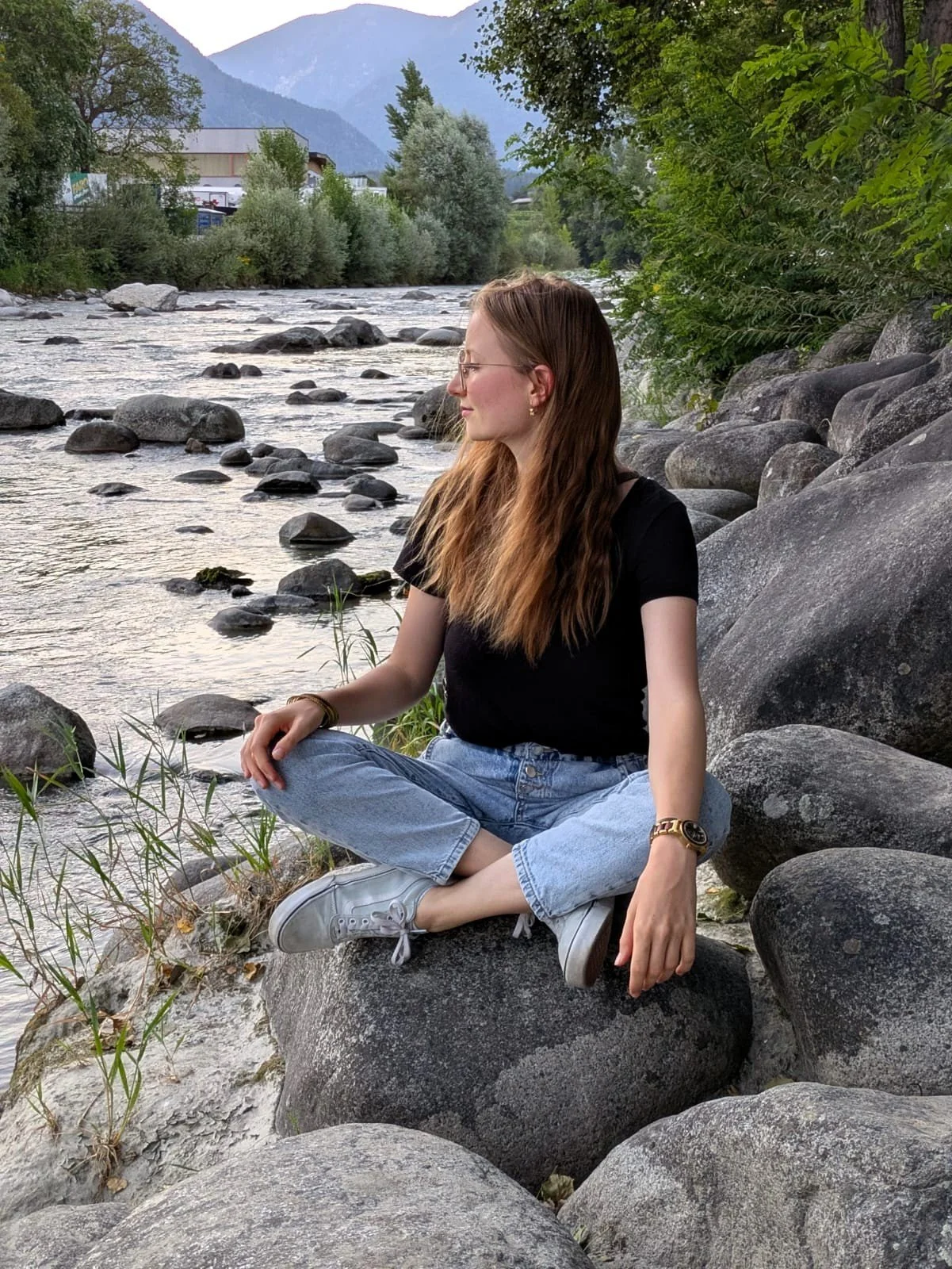 Eine junge Frau sitzt entspannt auf einem großen Stein am Fluss, umgeben von Steinen und Bäumen, mit Bergen im Hintergrund.