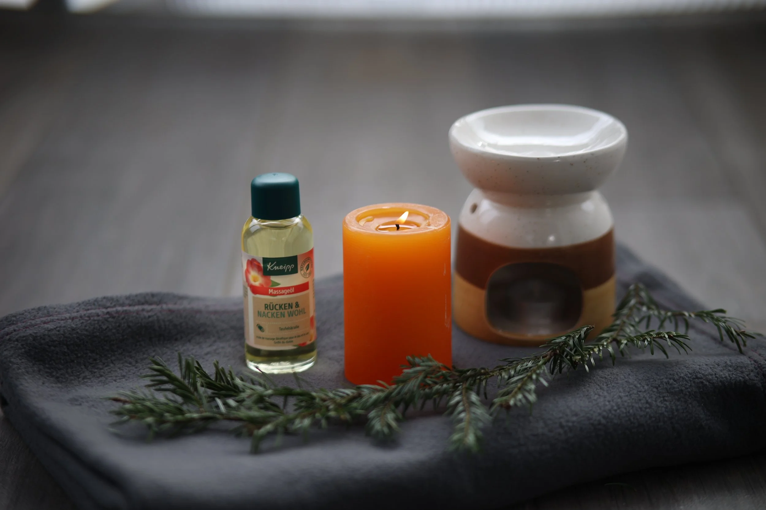 Spa-Arrangement mit einer weißen Duftlampe, einer brennenden Kerze, ätherischem Öl und Tannenzweigen auf einem grauen Handtuch.