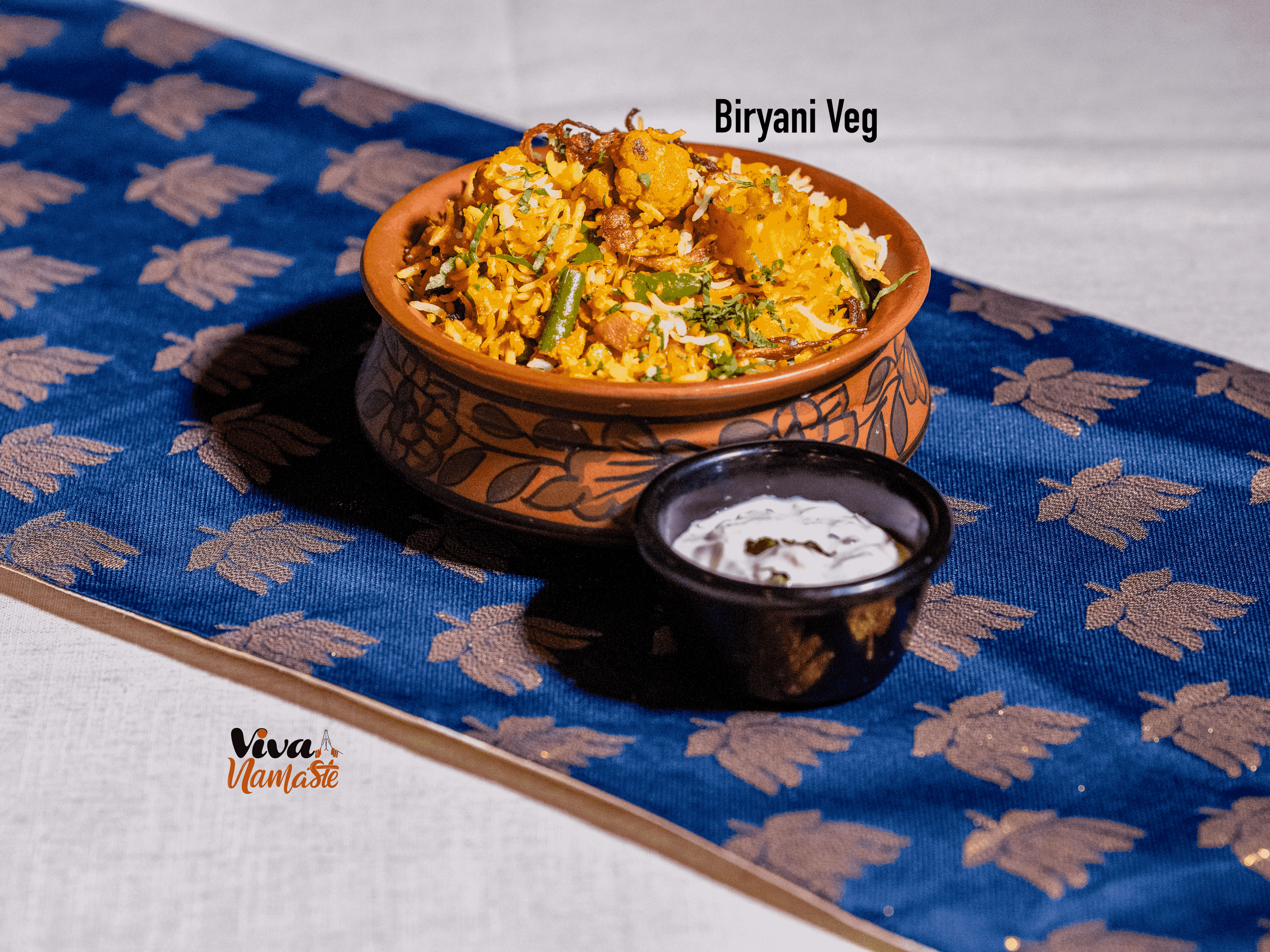 A bowl of vegetarian biryani rice with vegetables on a blue and gold patterned table runner, accompanied by a small black bowl of yogurt sauce that can be ordered from Viva Namaste—the Best Indian restaurant in Ashford