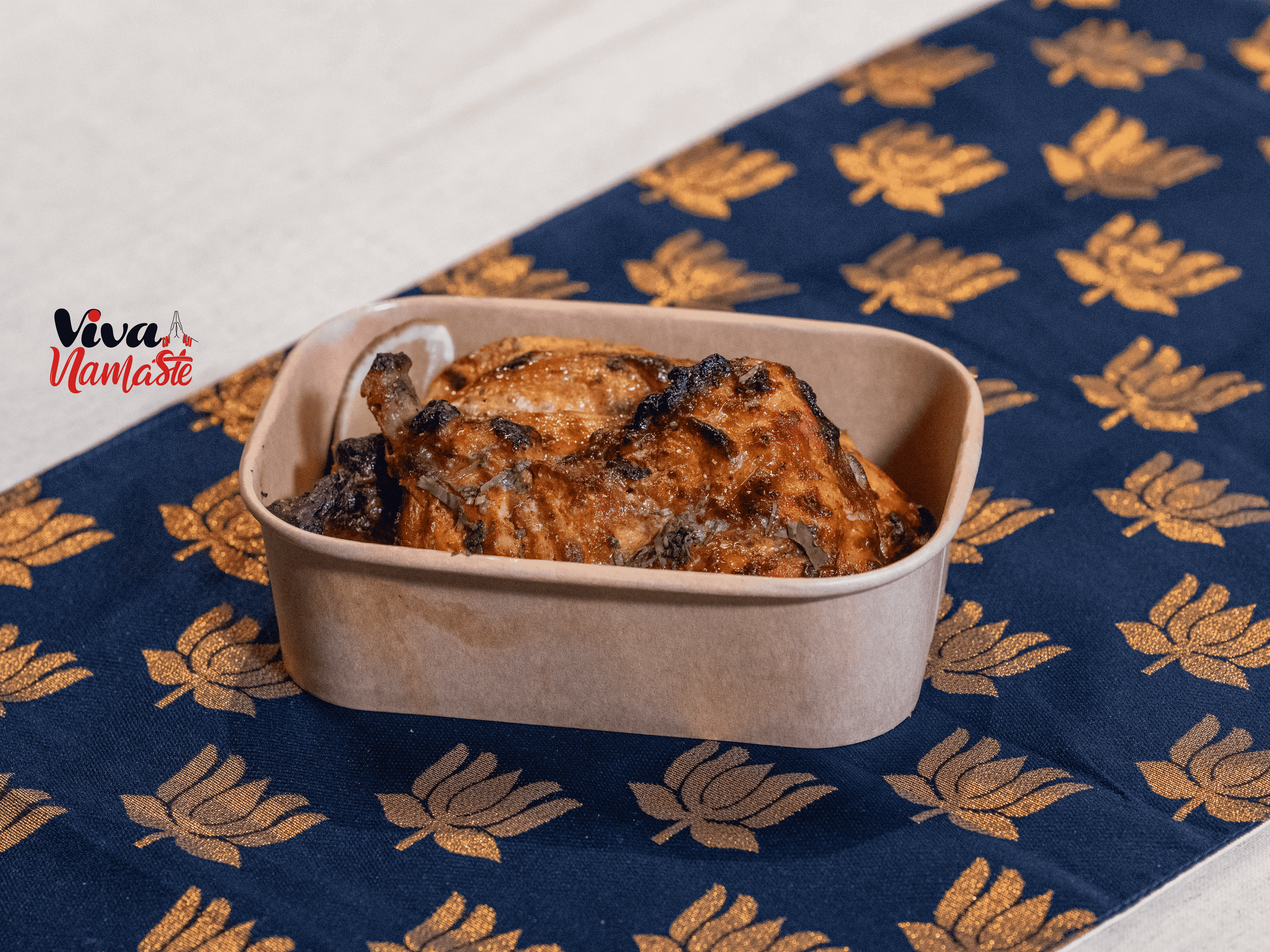 A baked chicken quarter in a brown container, placed on a blue cloth with golden floral patterns. Viva Namaste Available Box meals delivery Ashford