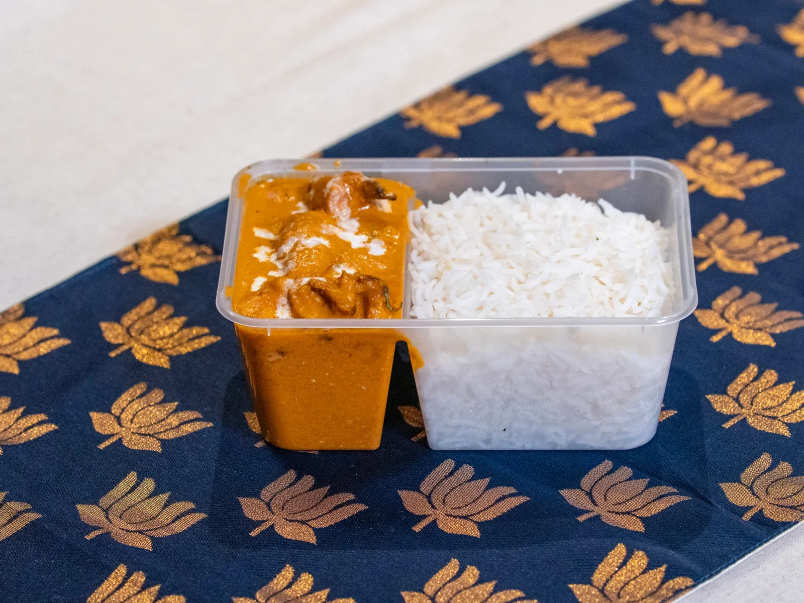 Home-style Indian meals in Ashford featuring rectangular transparent container with white rice on one side and butter chicken on the other, placed on a blue fabric with gold leaf patterns