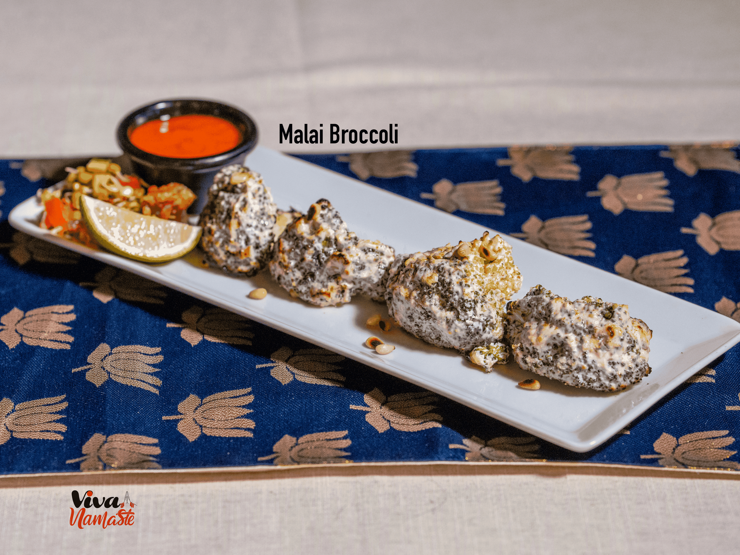 Plate of Malai broccoli, lemon wedge, and two types of chili or chutney from Viva Namaste Indian restaurant in Ashford with a small bowl of red sauce on a white rectangular plate, placed on a blue cloth with beige pattern.