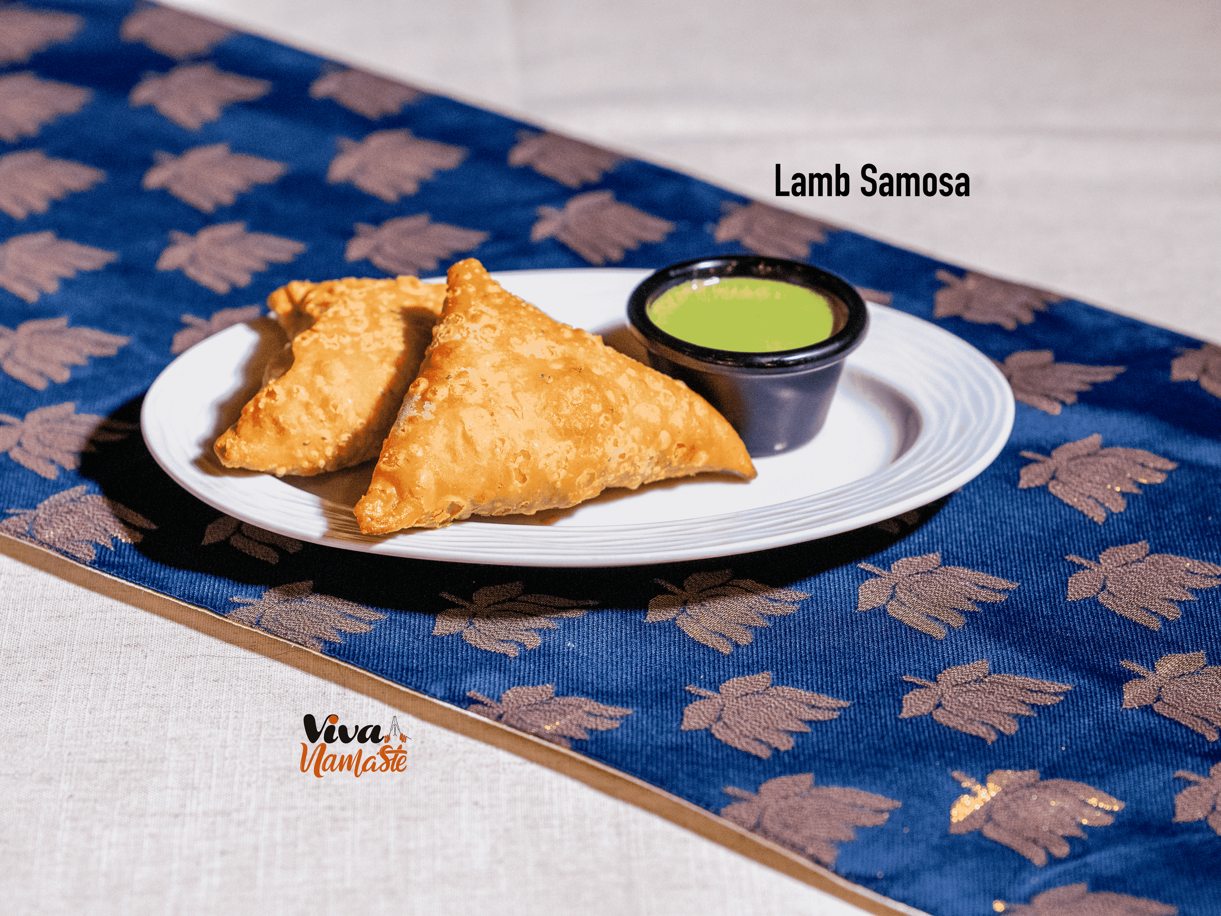 A white plate with two lamb samosas and a small container of green chutney, placed on a blue and brown decorative tablecloth from Viva Namaste Authentic Indian food near TW15
