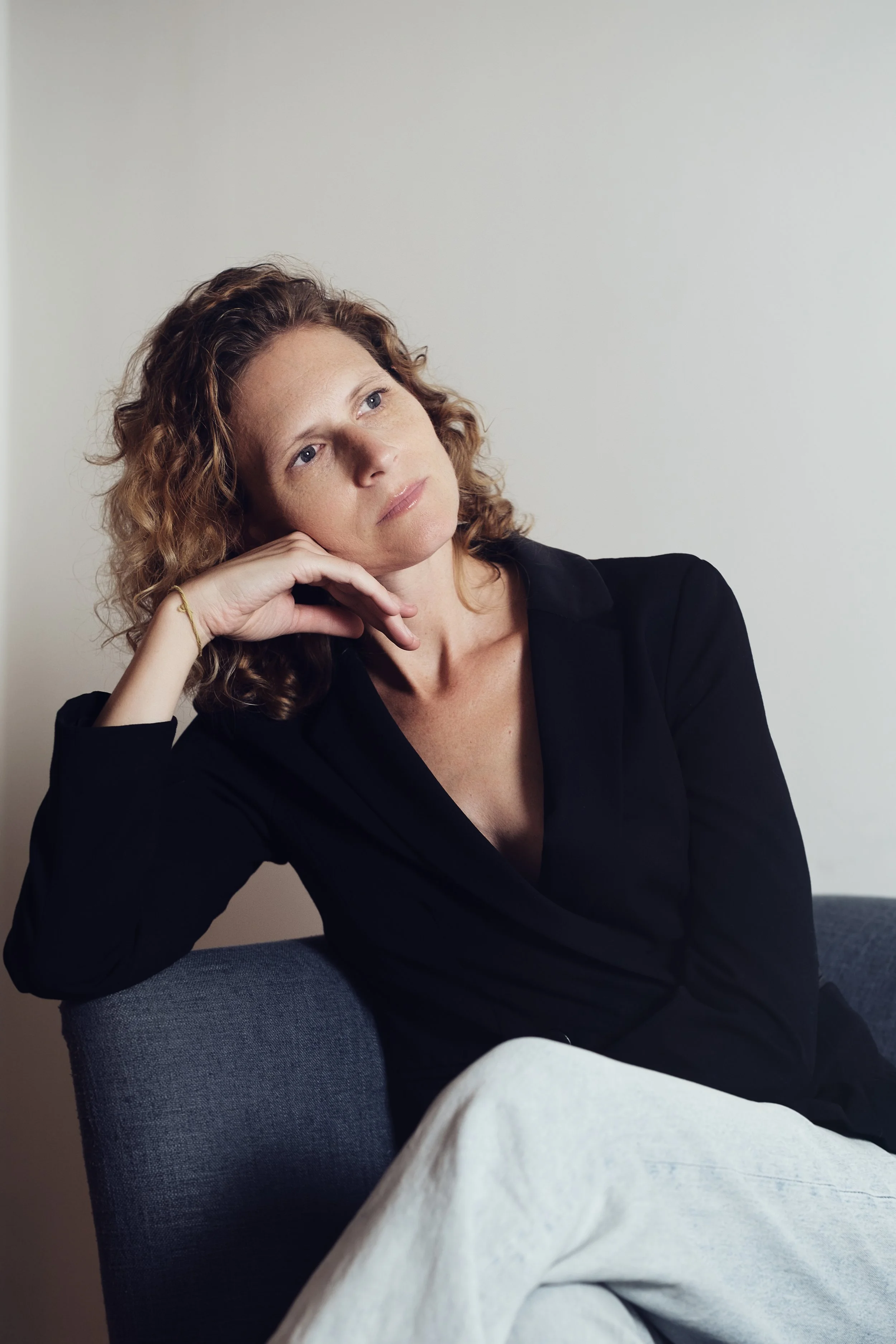 A woman with curly red hair sitting on a gray chair, resting her head on her hand, looking thoughtfully to the side against a plain white wall.
