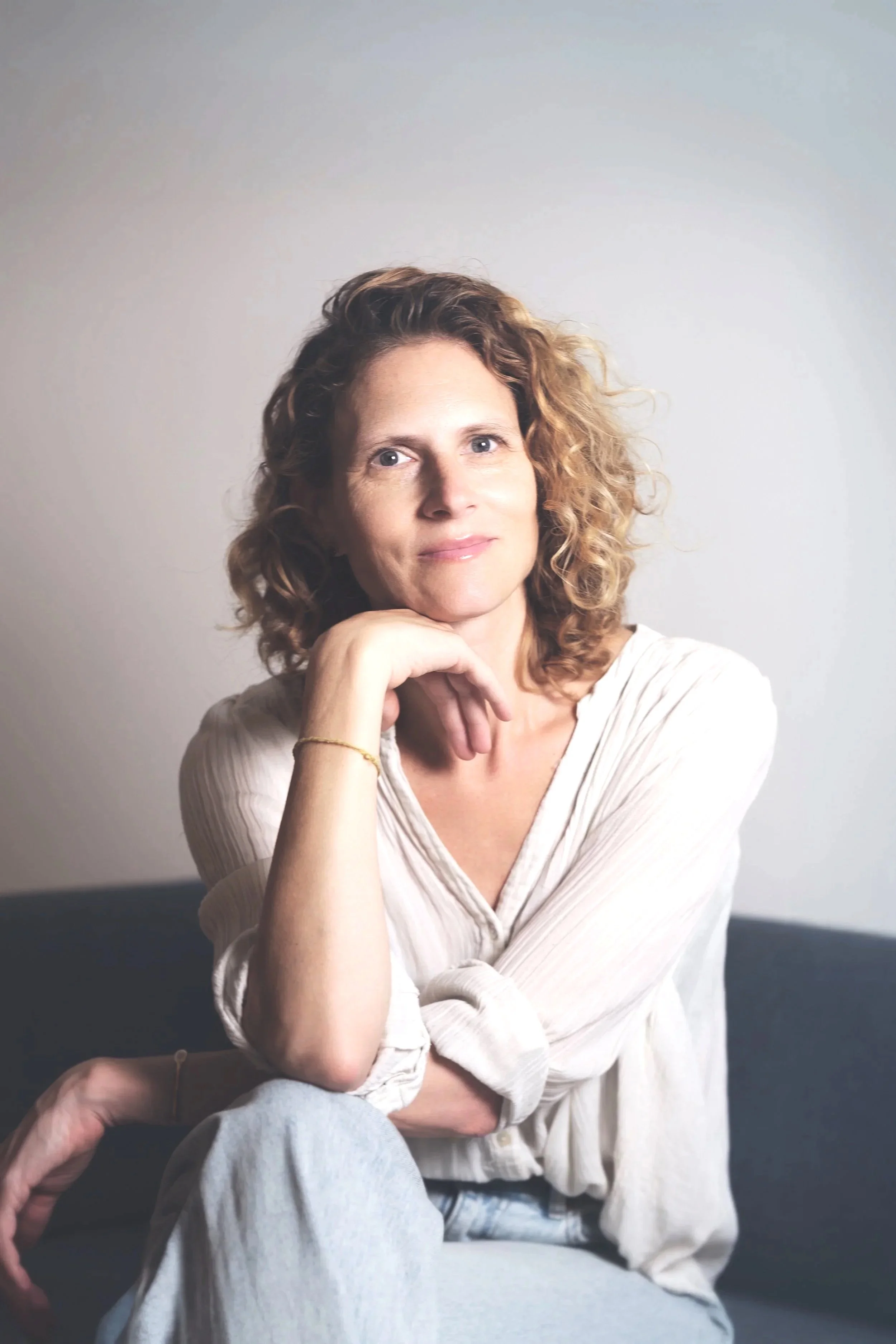 Portrait of a woman with curly hair sitting on a couch, resting her chin on her hand, wearing a white blouse and light-colored jeans, with a neutral background.