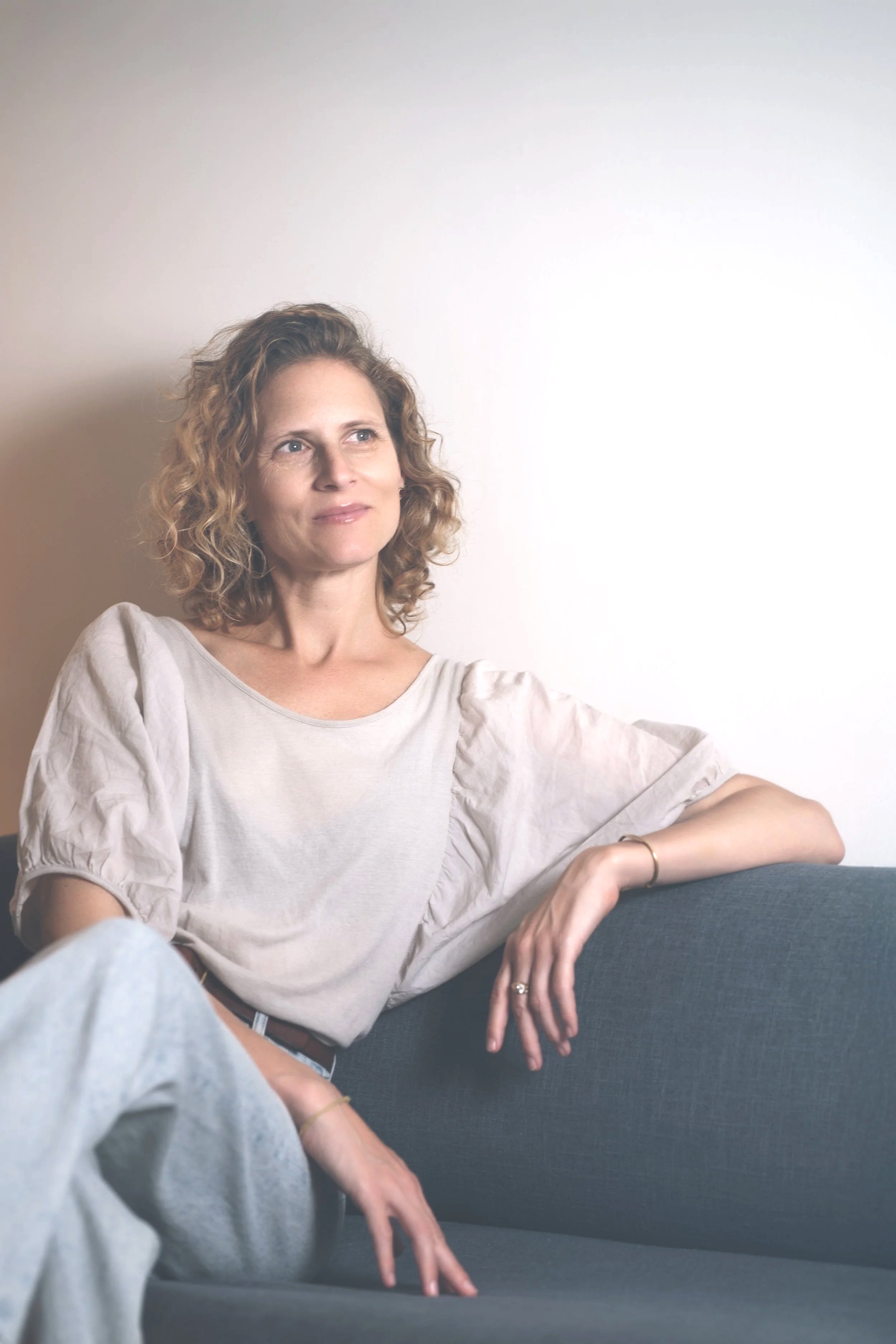 A woman with curly blonde hair sitting on a dark gray couch against a plain white wall. She is wearing a light beige top with puffed sleeves and light-colored pants, looking thoughtfully to her right.