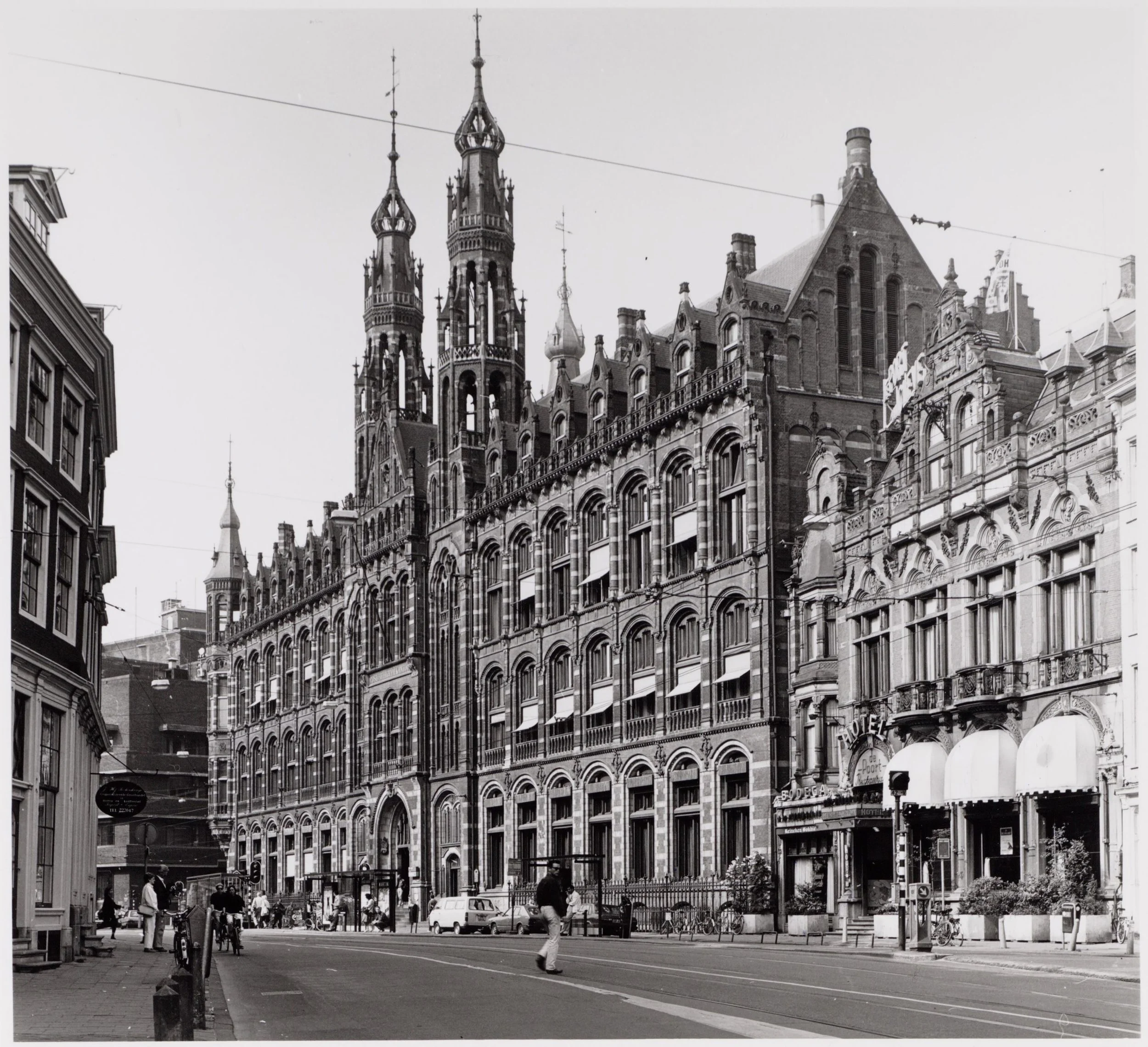 Black and white photo of historic European-style buildings on a city street, with people walking and bicycles parked, and a hotel sign visible.