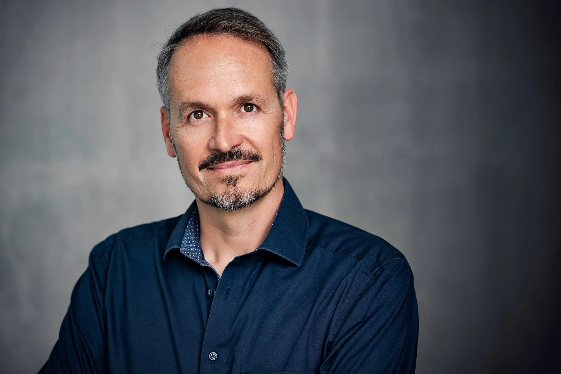 Close-up portrait of a middle-aged man with short gray hair, a goatee, and mustache, wearing a dark blue button-up shirt, against a blurred gray background.