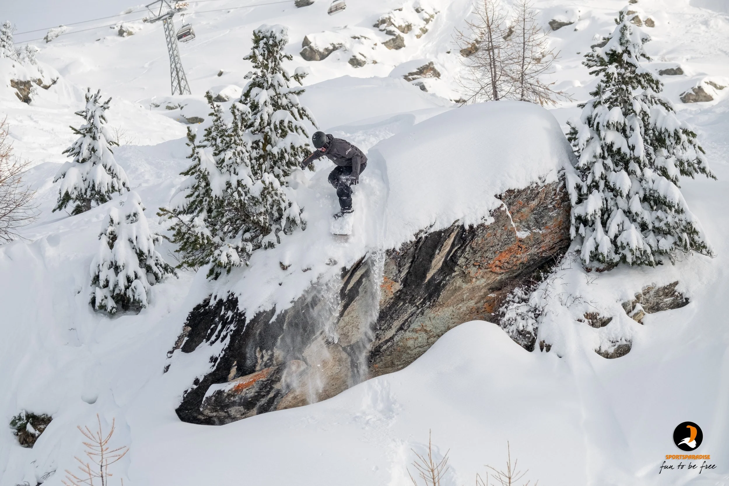 A snowboarder in black gear snowboarding off a large snow-covered rock, surrounded by snow-covered trees in a winter landscape.