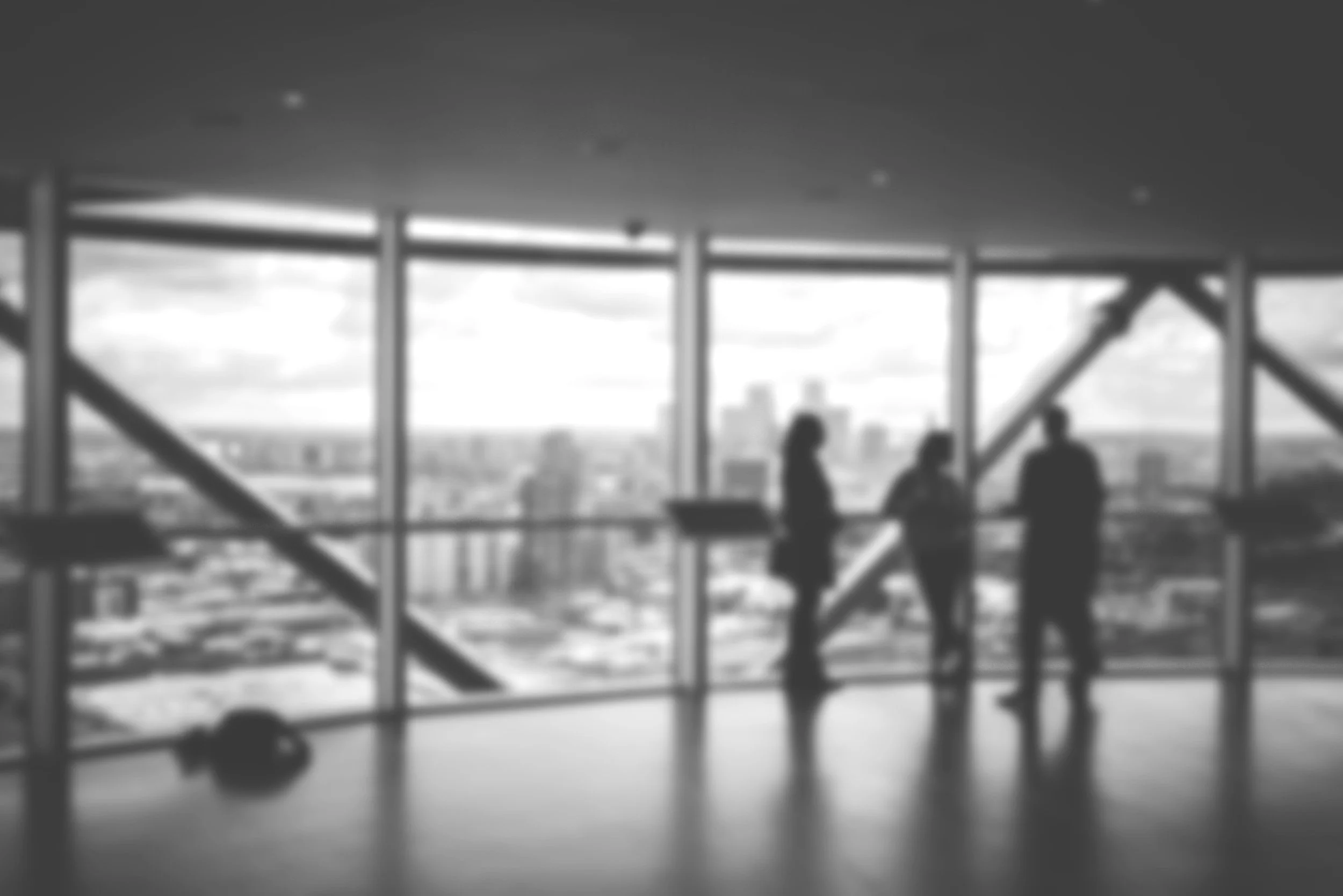 Black and white photo of people sitting inside a building with large windows, overlooking a cityscape, some standing and some sitting, with visible structural beams.