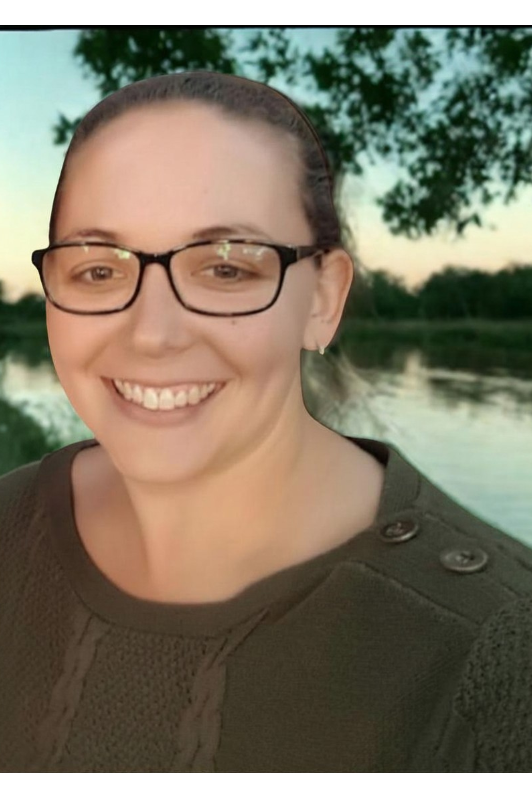 A woman with glasses and earrings smiling outdoors with a lake and trees in the background at sunset or sunrise.