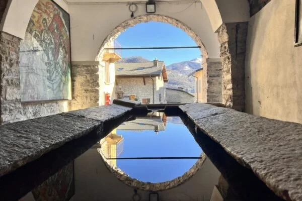 Una piazza con un ristorante all'aperto, tavoli con sedie e ombrelloni, una fontana in primo piano e un edificio colorato con balconi e finestre.