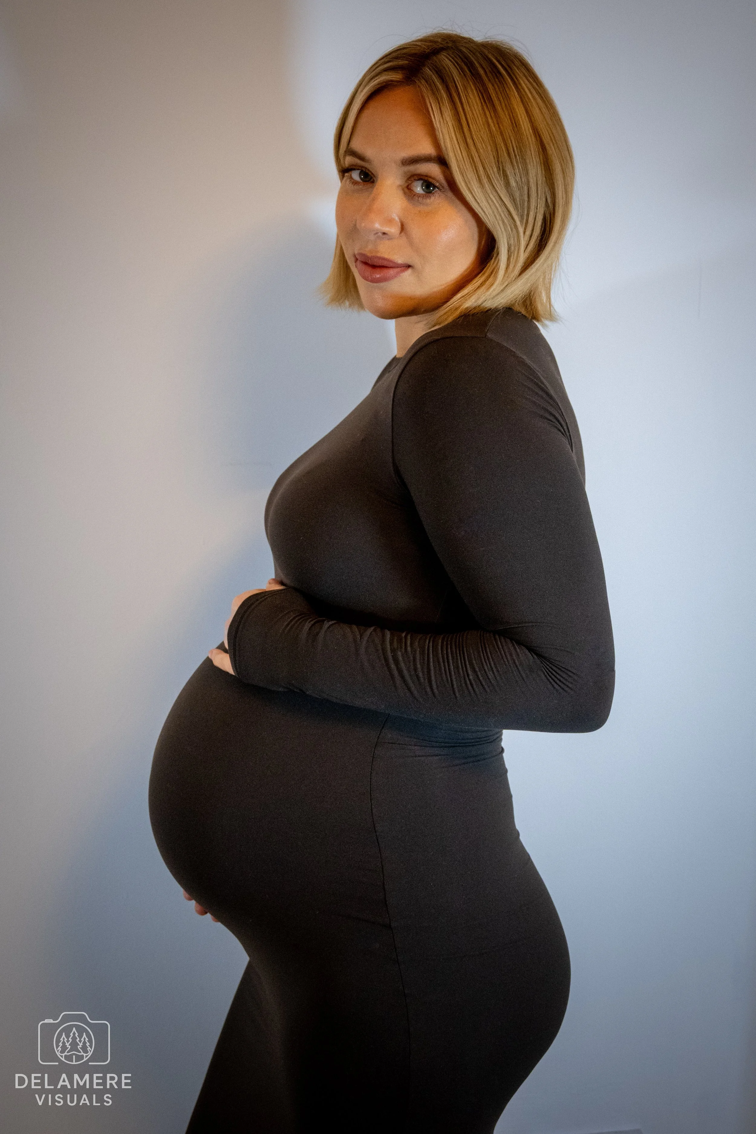 A pregnant woman with short blonde hair looking at the camera and smiling. She is wearing a long, fitted black dress and her hands are on her baby bump. Taken in Cheshire.