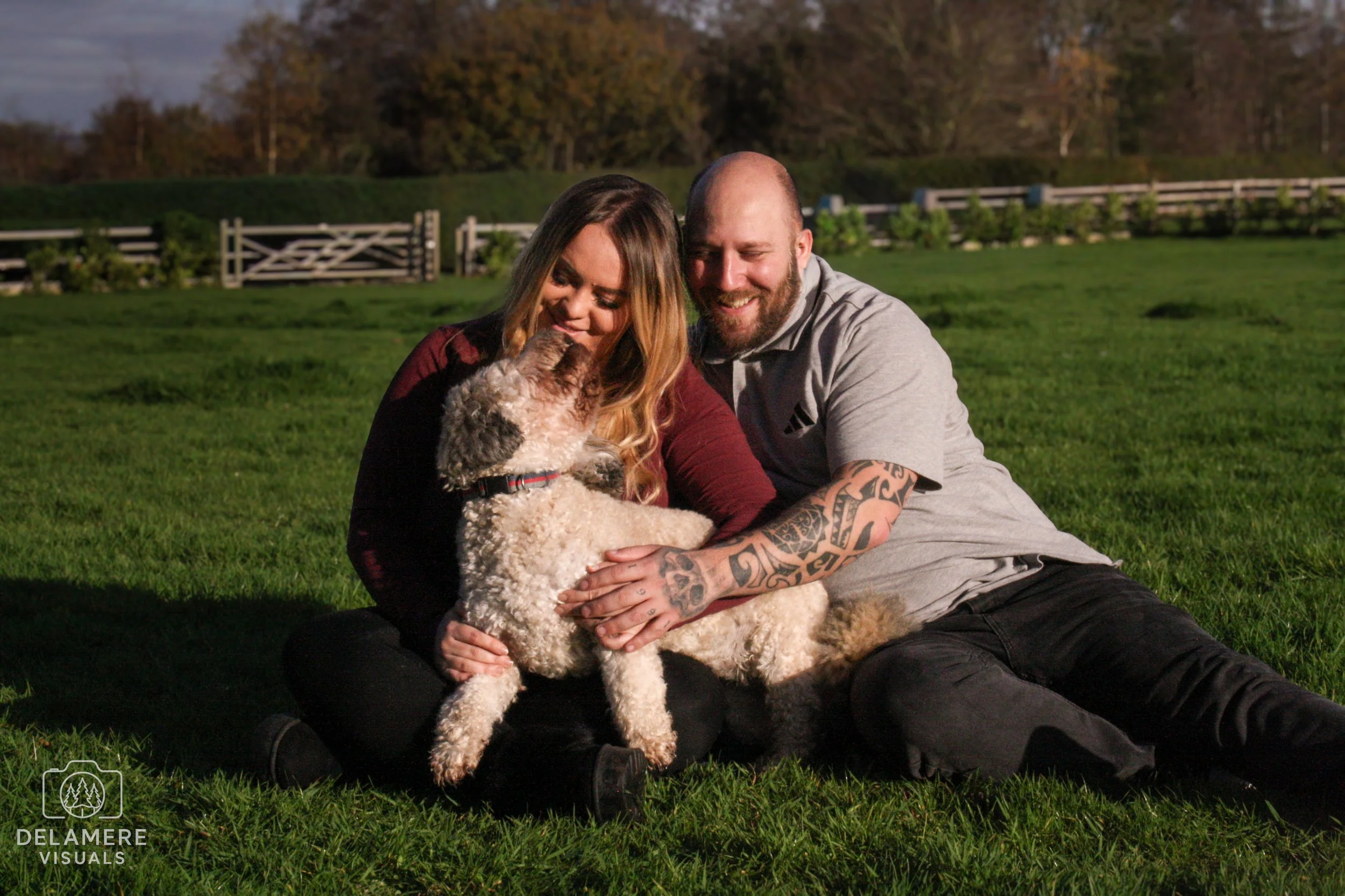 A happy couple sitting on the grass with their dog, kissing and smiling outdoors during daytime. Taken in Cheshire.