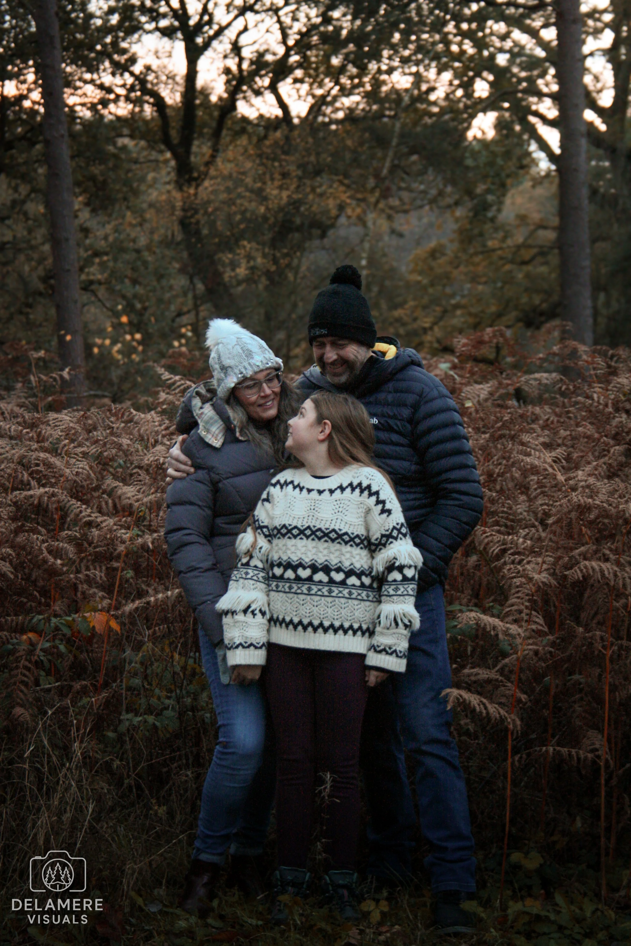 A family of three standing close together outdoors in a forest, smiling and looking at each other, wearing winter clothing, with trees and autumn foliage in the background. Taken in Cheshire.