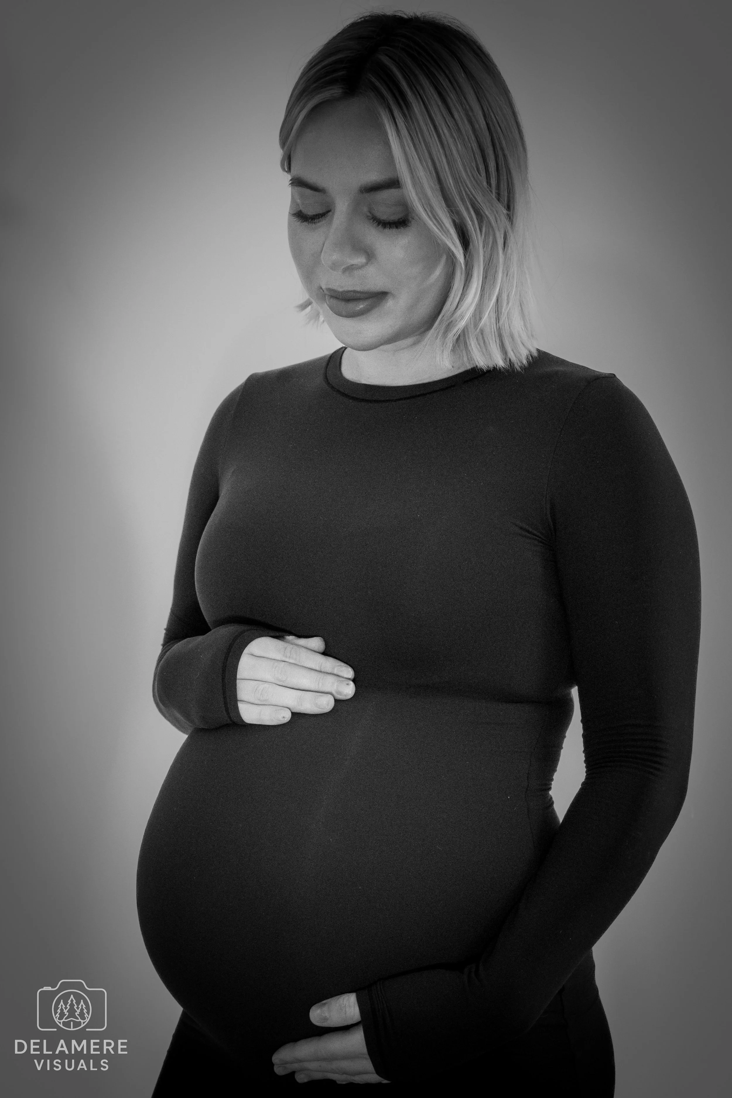 A pregnant woman with short blonde hair looking at the camera and smiling. She is wearing a long, fitted black dress and her hands are on her baby bump. Taken in Cheshire.
