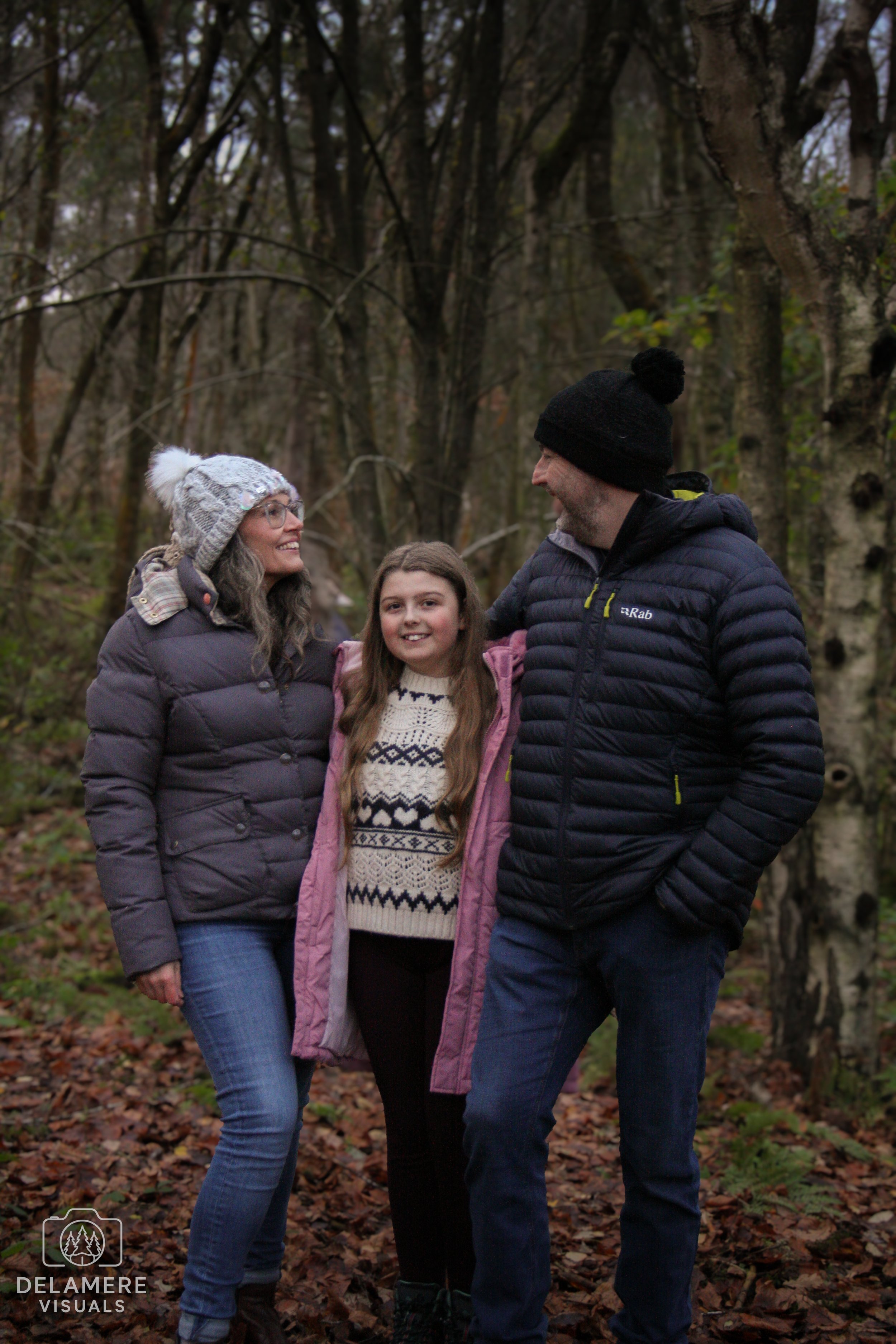 A family of three, two adults and a girl, smiling and walking outdoors in a wooded area during fall, dressed in warm jackets and hats. Taken in Cheshire.