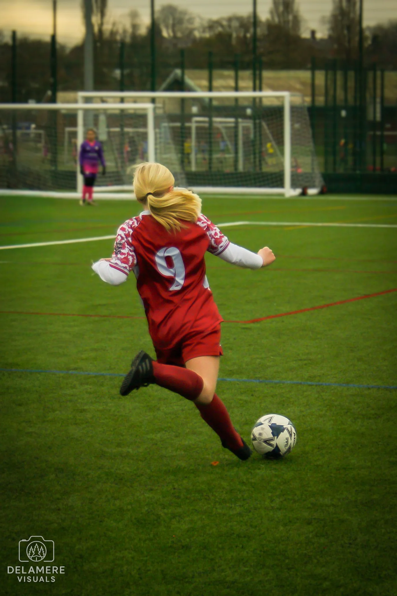 A girl in a football uniform kicking a football on a field during daylight, with a goalie and other players in the background. Taken in Cheshire.