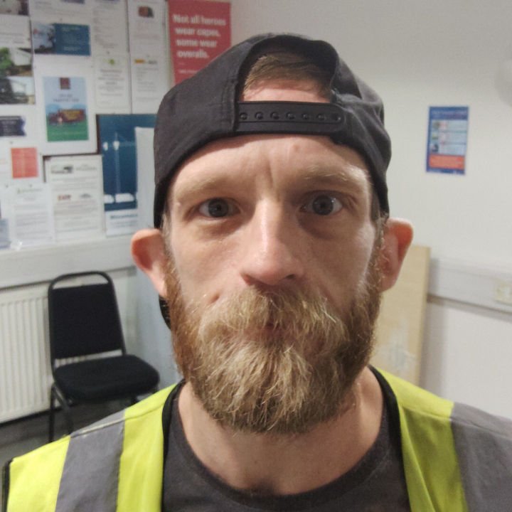 A man with a beard wearing a backward black cap and neon safety vest, looking at the camera indoors with posters and a chair in the background.