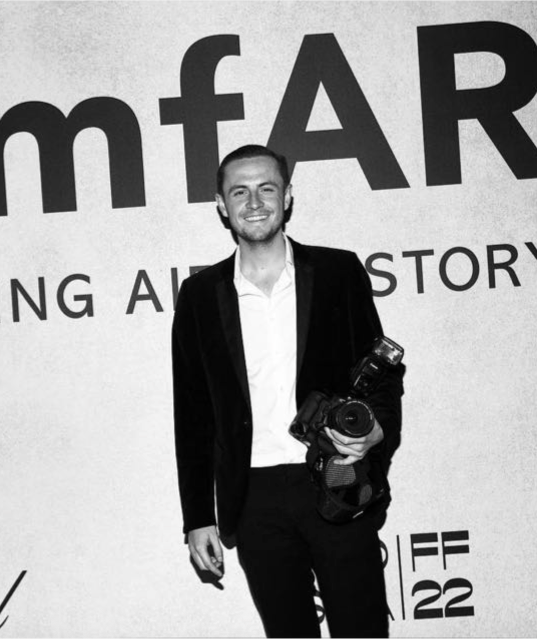 A man smiling while holding a professional camera in front of a large sign with the word 'mear' in bold letters.