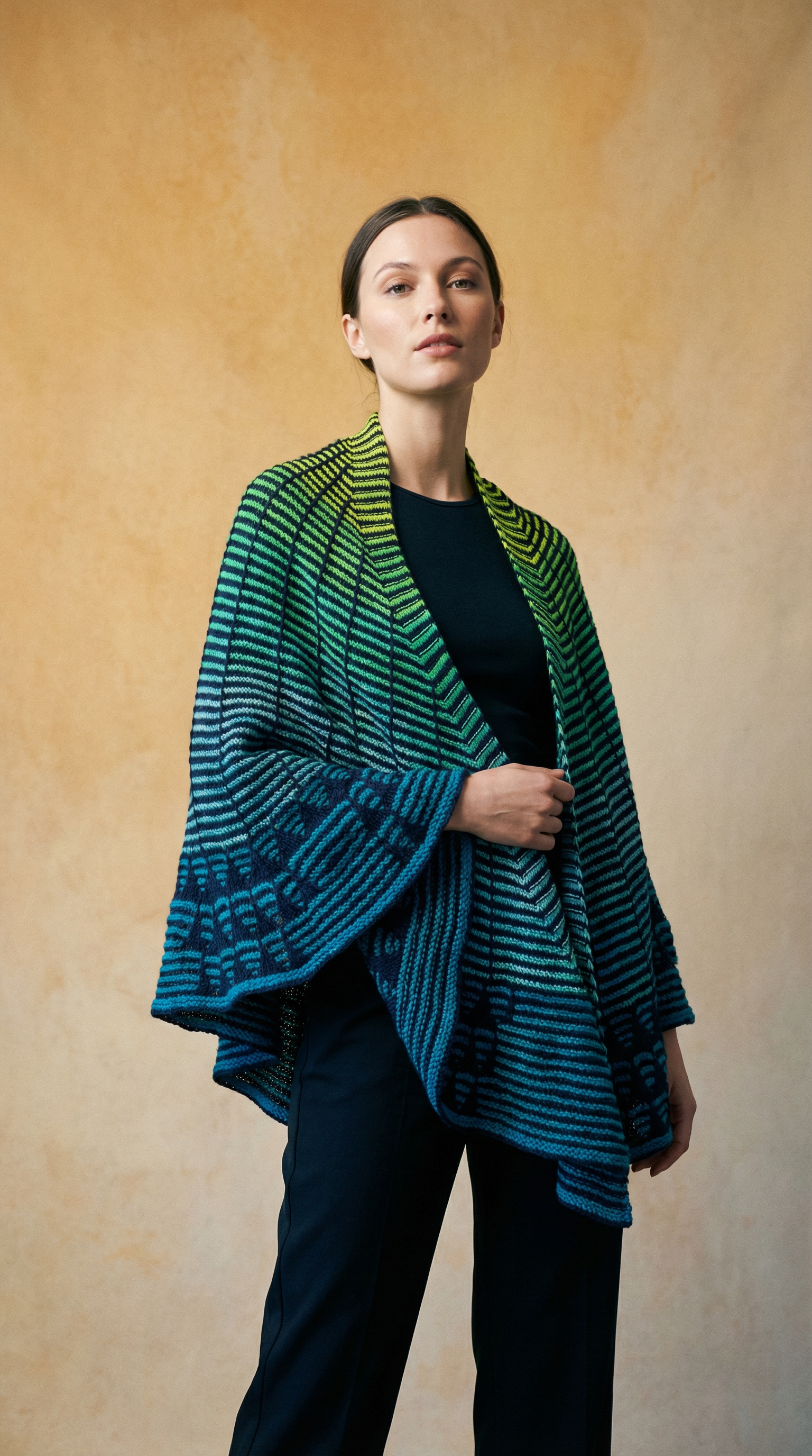 A woman wearing a black top and black pants, with a colorful, knitted shawl, stands against a textured beige background.