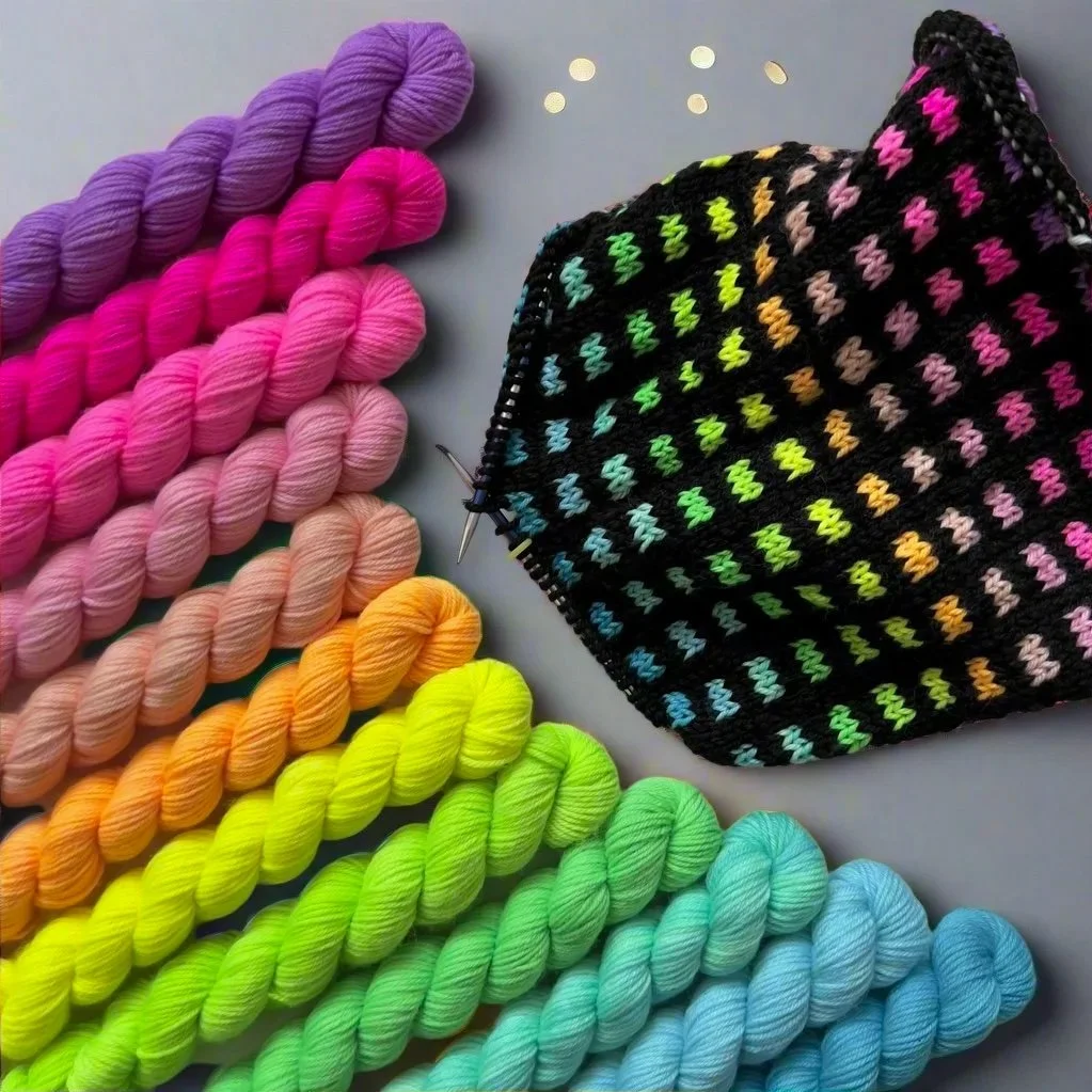 Colorful skeins of yarn in purple, pink, peach, yellow, green, and blue, along with a partially completed black knitted cowl with rainbow-colored stitches.
