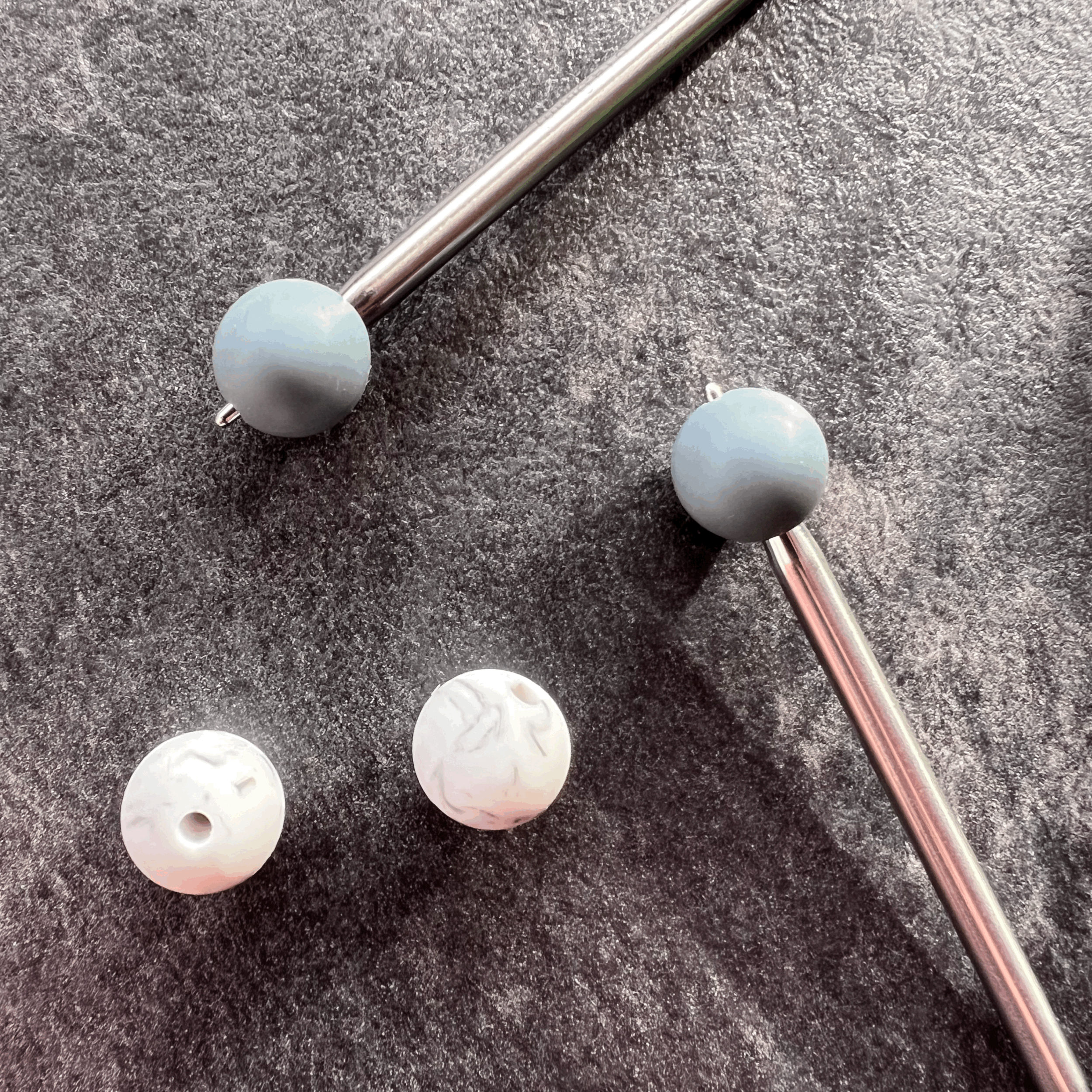 Two metallic knitting needles with white and grey needle stoppers, sitting on a textured grey surface.