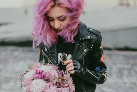 An comtemplative, artistic-looking young woman with pink hair holding a bouquet of pink flowers outdoors.