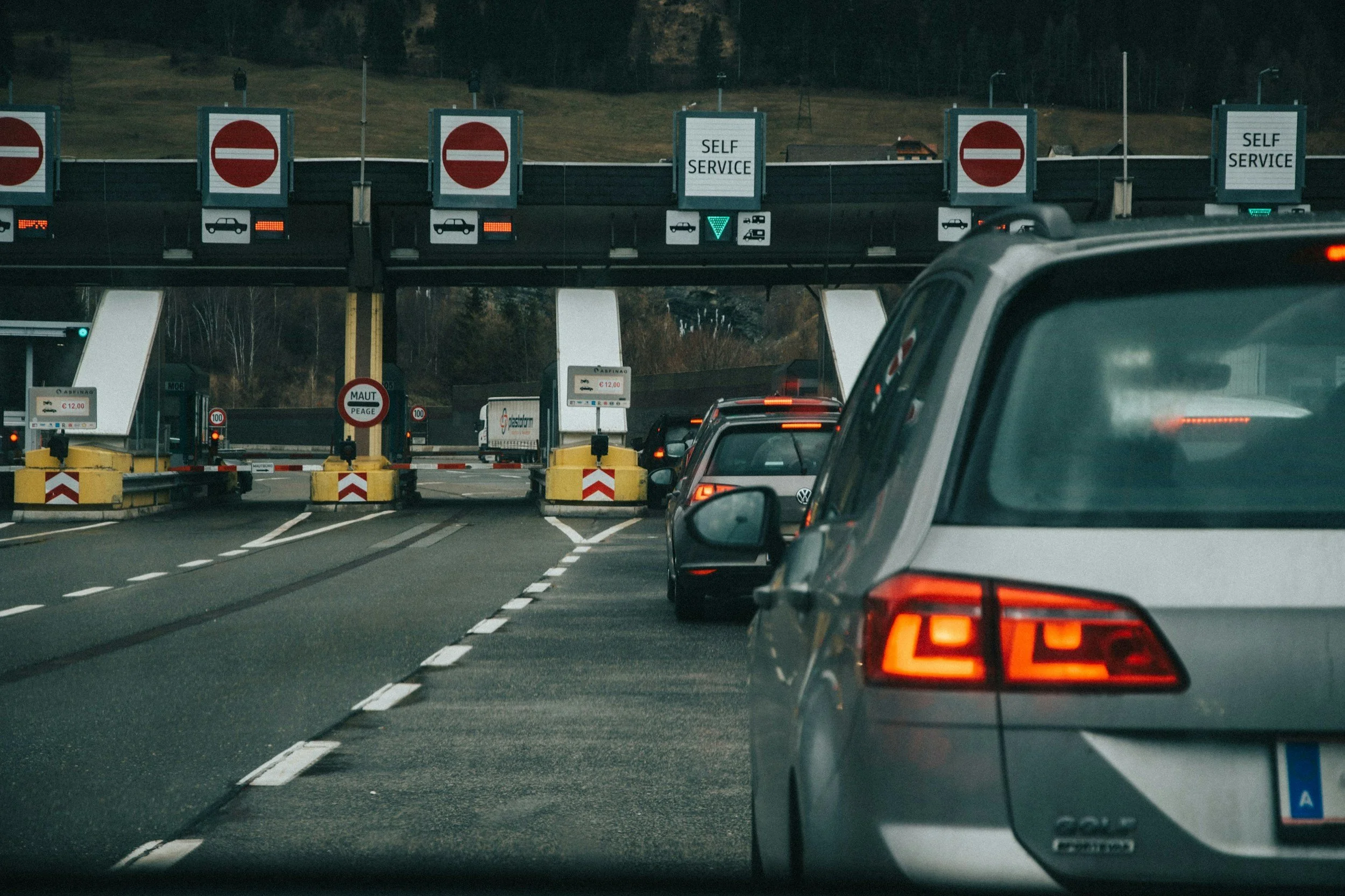 Line of cars passing through a toll booth with signs indicating 'Self Service' in the background.