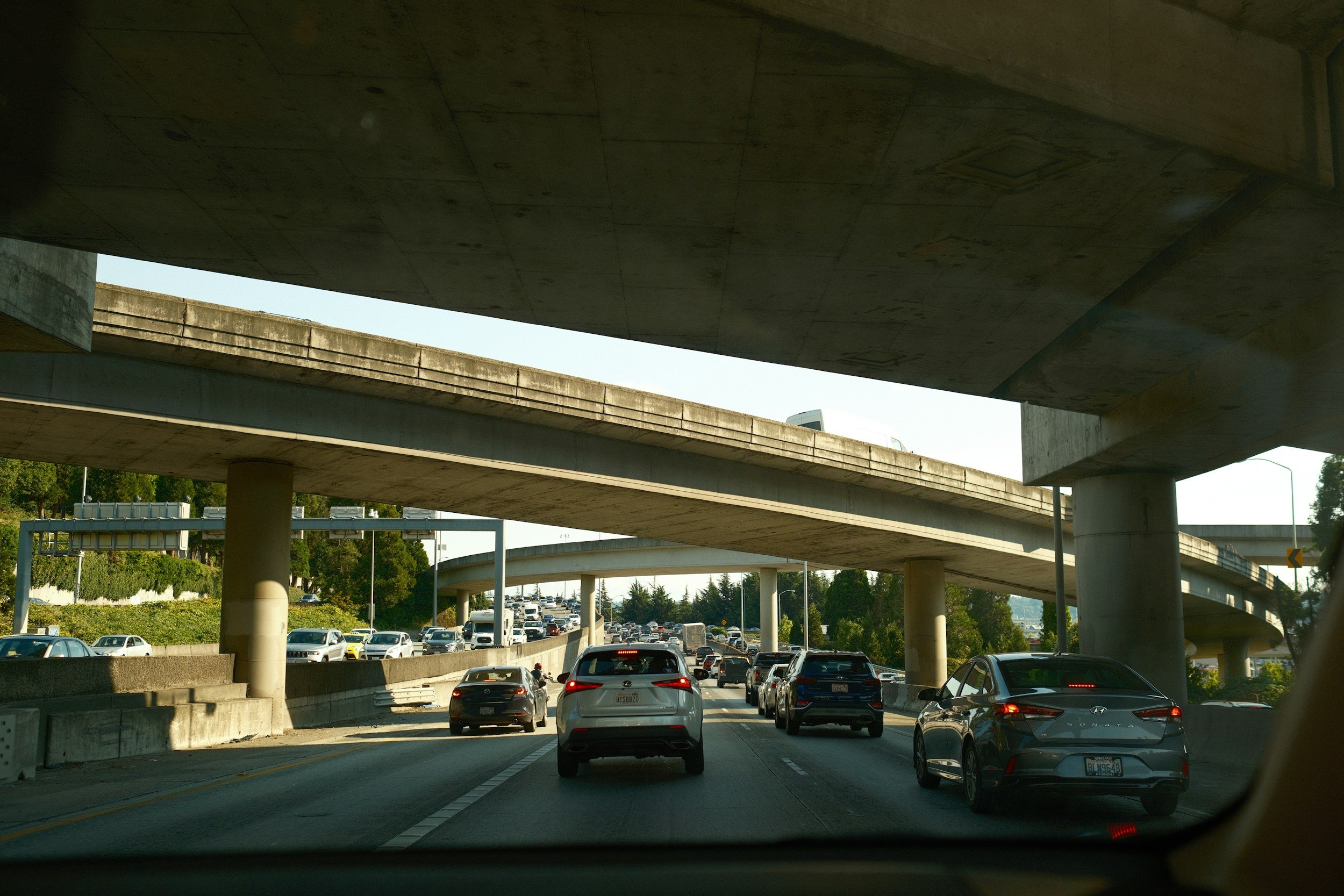 Traffic congestion on a multi-level highway, viewed from inside a vehicle. Several cars are seen on the road beneath and above the overpasses, with trees and greenery in the background.