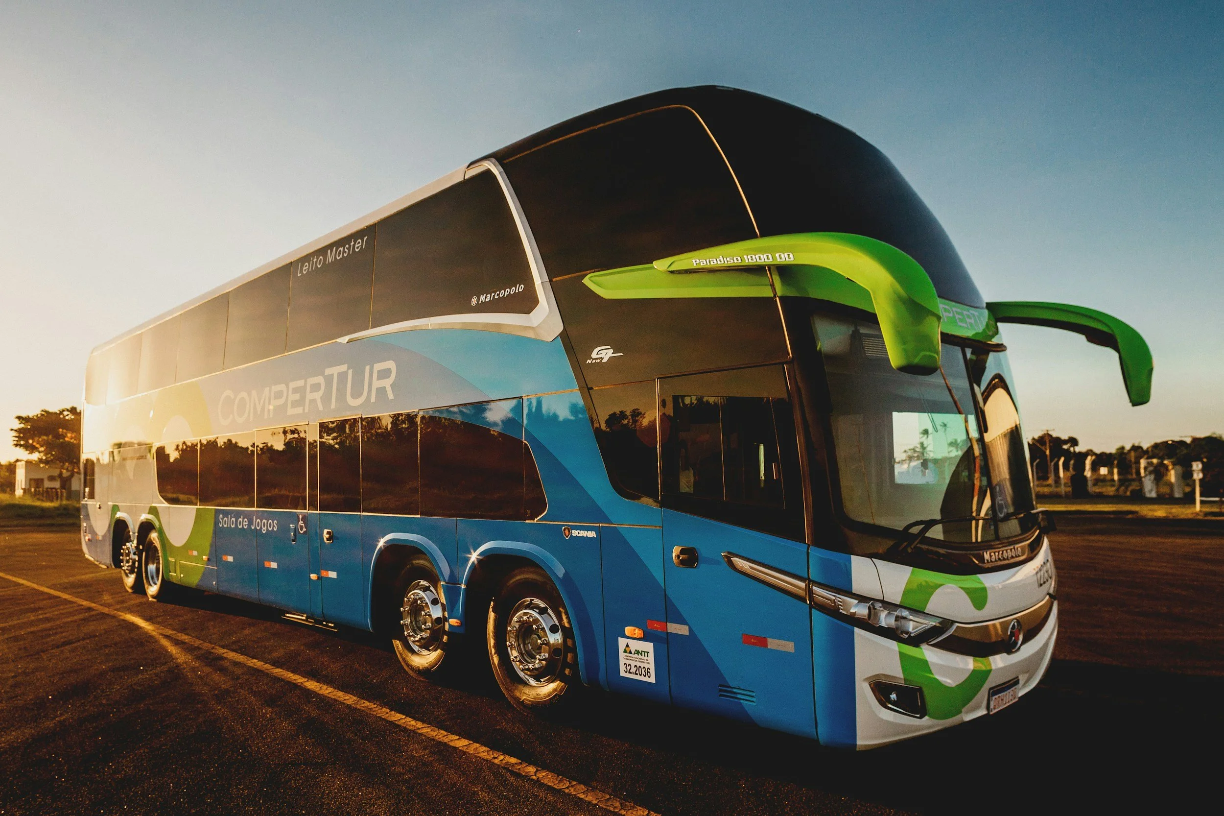 A modern double-decker bus parked on a street at sunset, painted in blue, white, and green with branding.