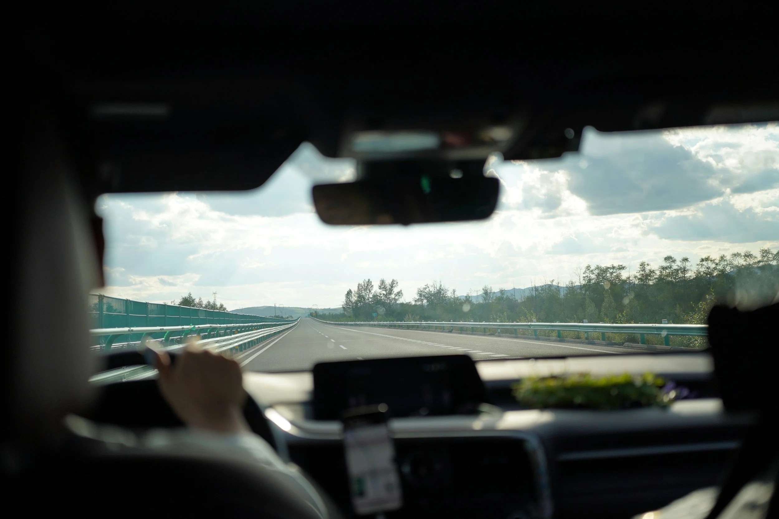 Inside a vehicle, view from the back seat looking out at an empty highway with guardrails and a cloudy sky.