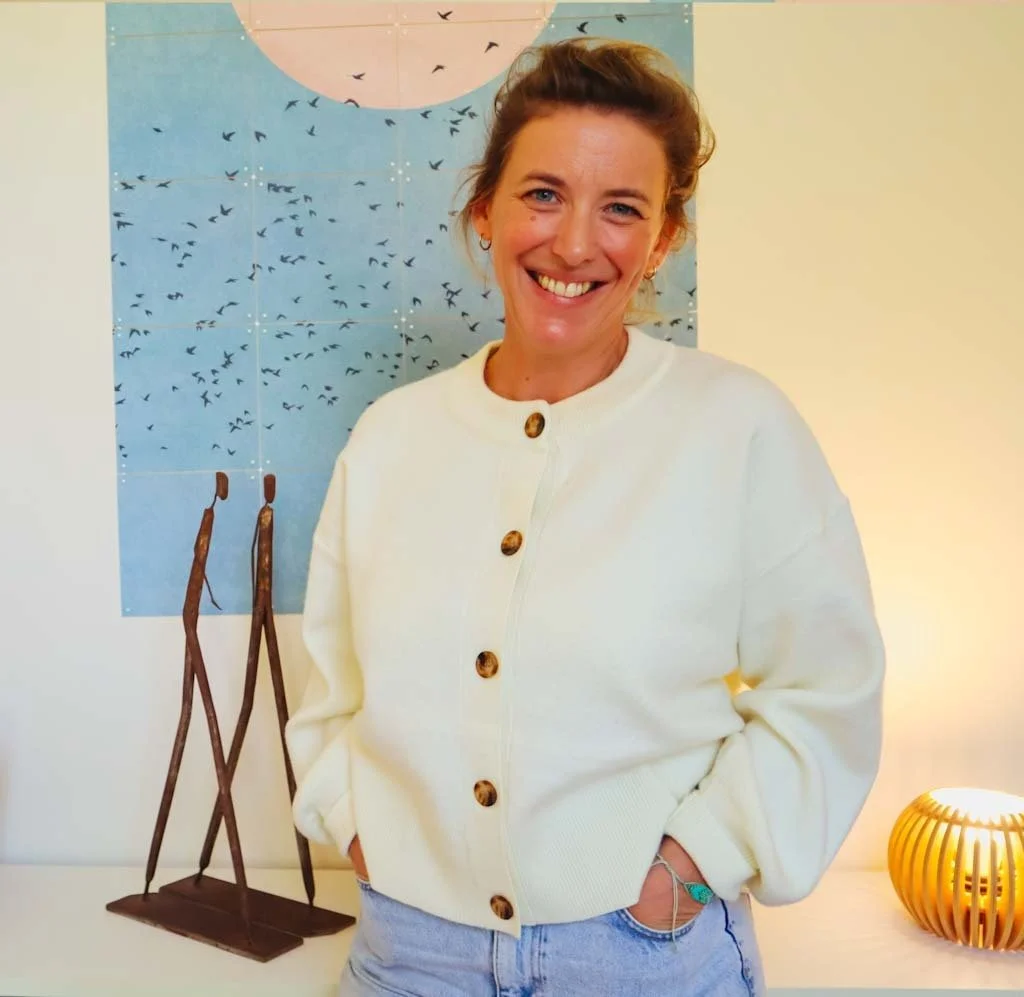 Une femme souriante, portant un pull blanc à boutons, se trouve devant un tableau, avec une décoration en bois et une lampe moderne à droite.