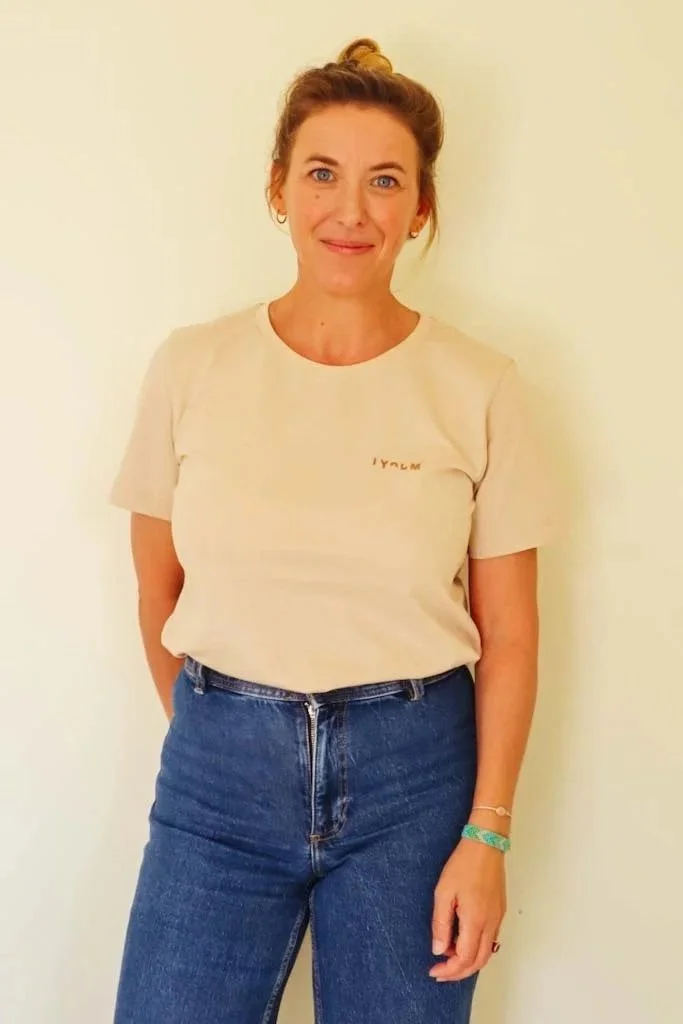 Une femme debout contre un mur blanc, portant un t-shirt beige et un jean bleu, avec des boucles d'oreilles et un bracelet.
