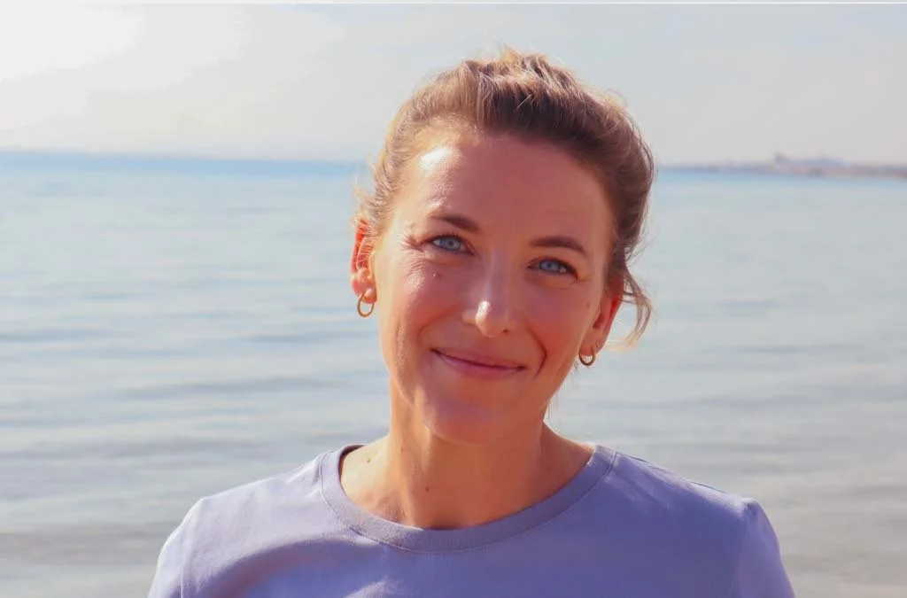 Une femme souriante avec des cheveux marron, portant des boucles d'oreilles  et un t-shirt violet, se tient près de la mer avec un ciel clair en arrière-plan.