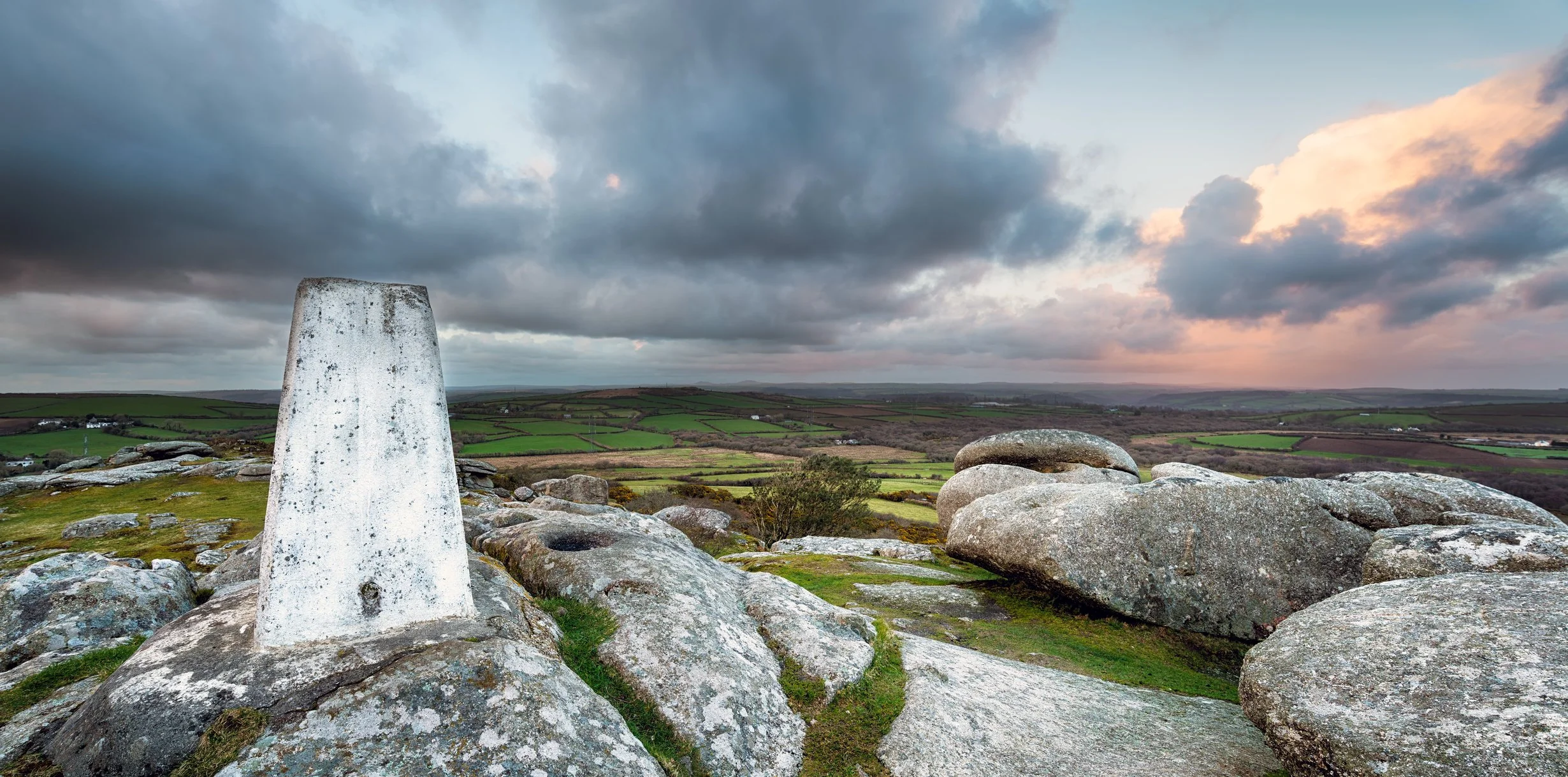 View of ancient rock formations and a stone marker on a hilltop overlooking a patchwork of green fields under a cloudy sky at sunset.