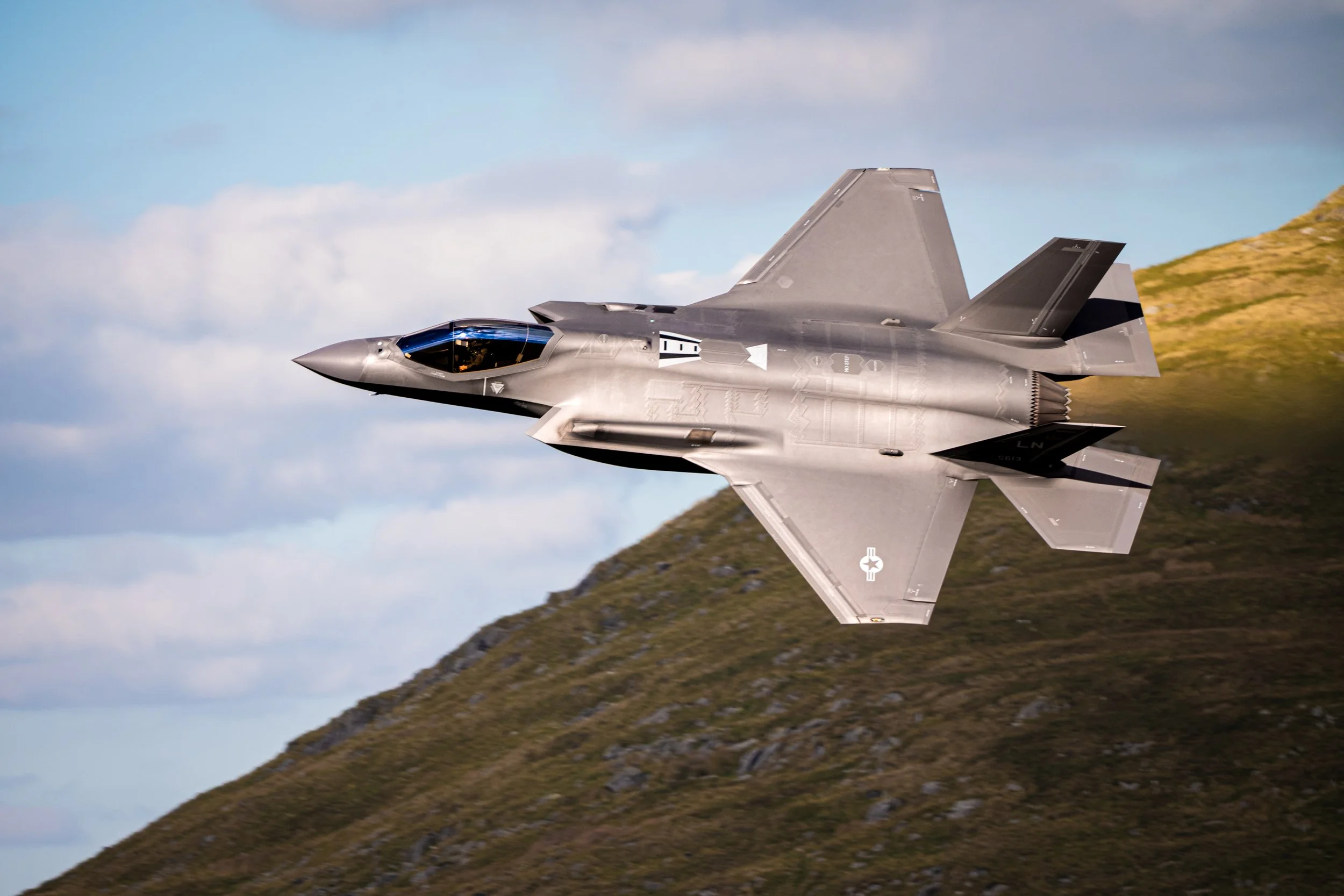 A fighter jet flying over a mountain landscape during the daytime.