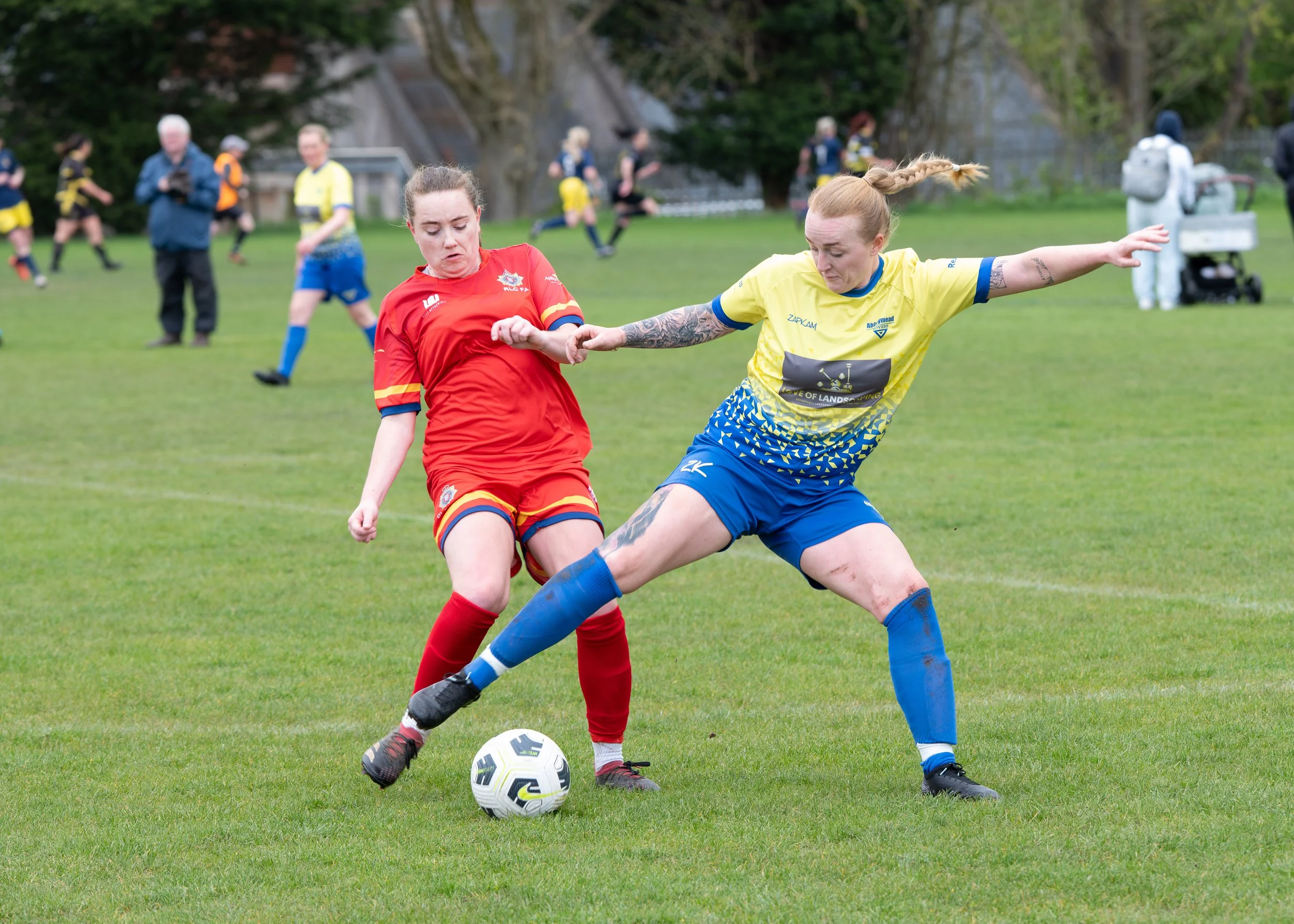 Two women playing soccer, one in a red uniform and the other in a yellow and blue uniform, competing for the ball on a grassy field with other players and spectators in the background.