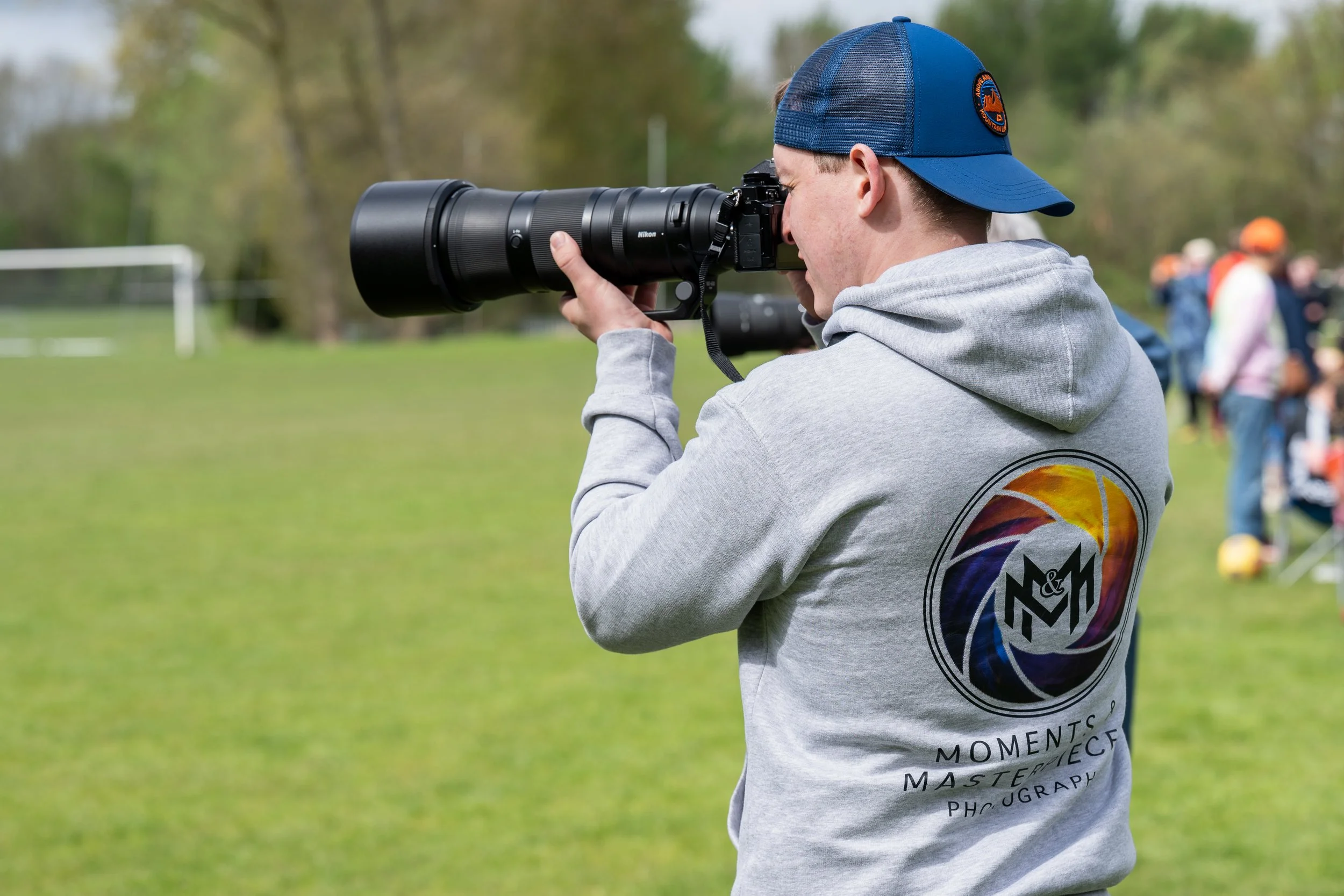 A person wearing a blue cap and a gray hoodie with printed text on the back, aiming a Nikon camera with a large telephoto lens at an outdoor event on a grassy field. In the background, there are blurred people and trees.
