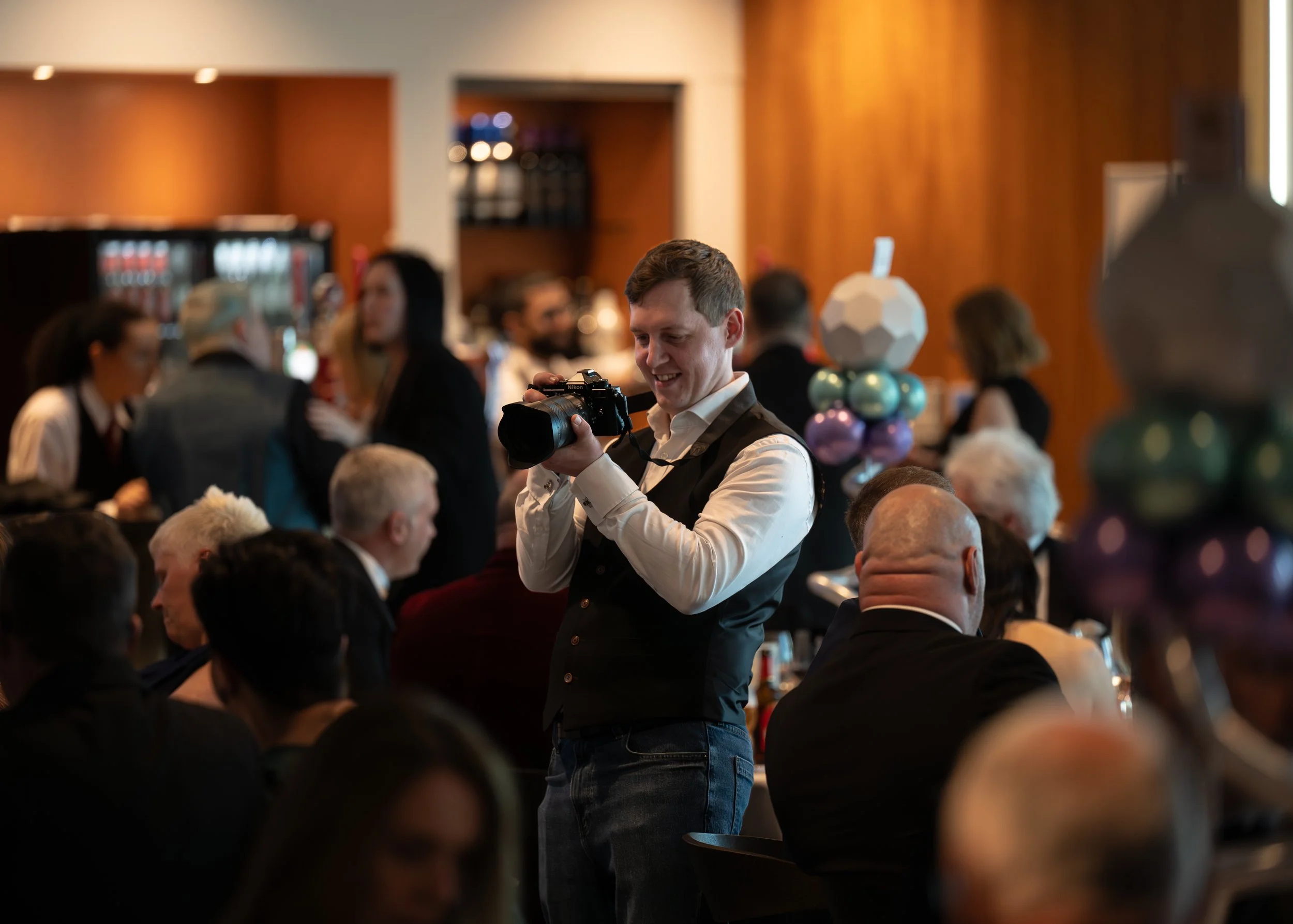 A smiling man in formal attire holding a camera at a crowded indoor event with people seated and standing, and decorative balloons on the table.