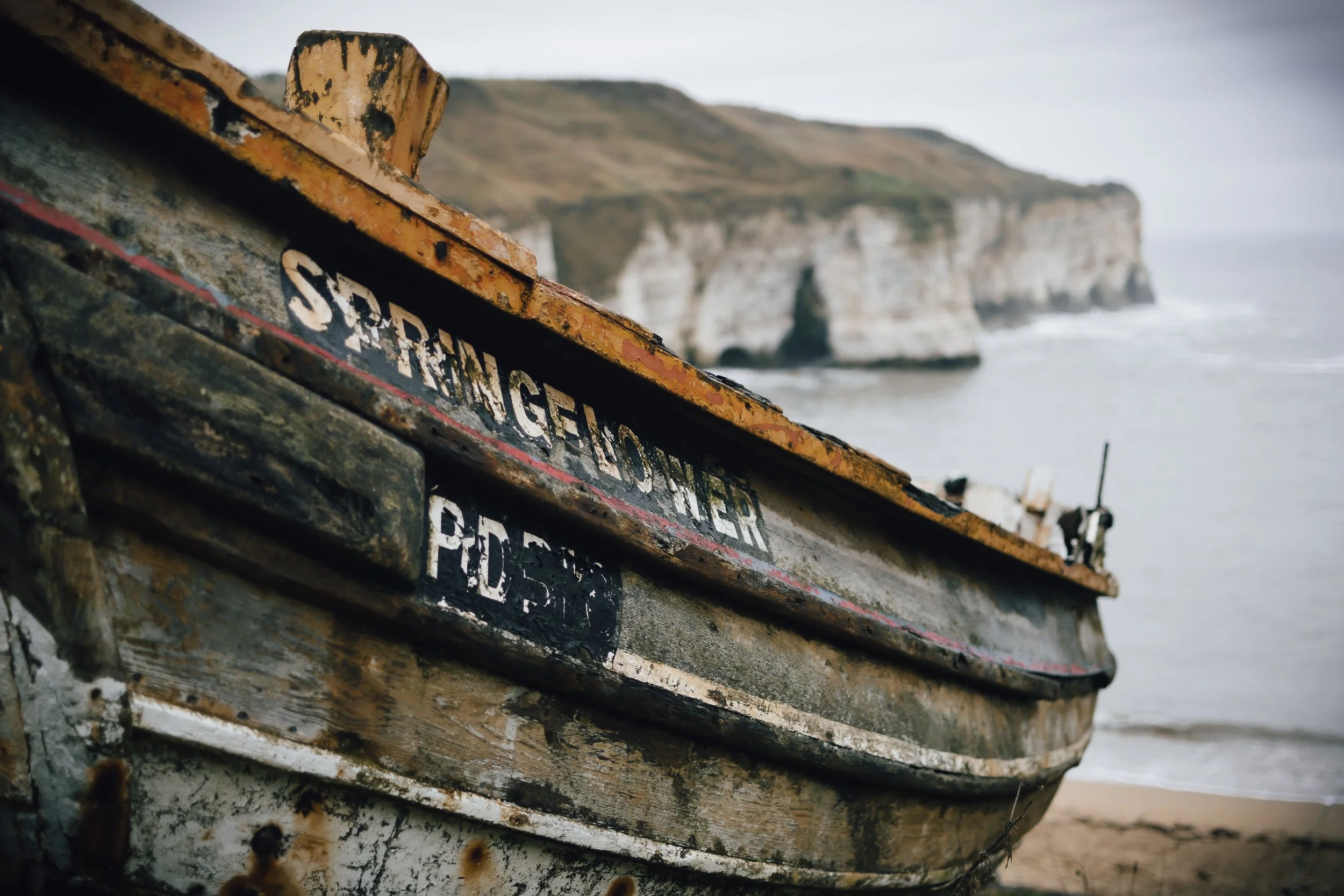 Close-up of an old, weathered, yellow and black boat with chipped paint, resting on the beach near the cliffs and ocean under an overcast sky.